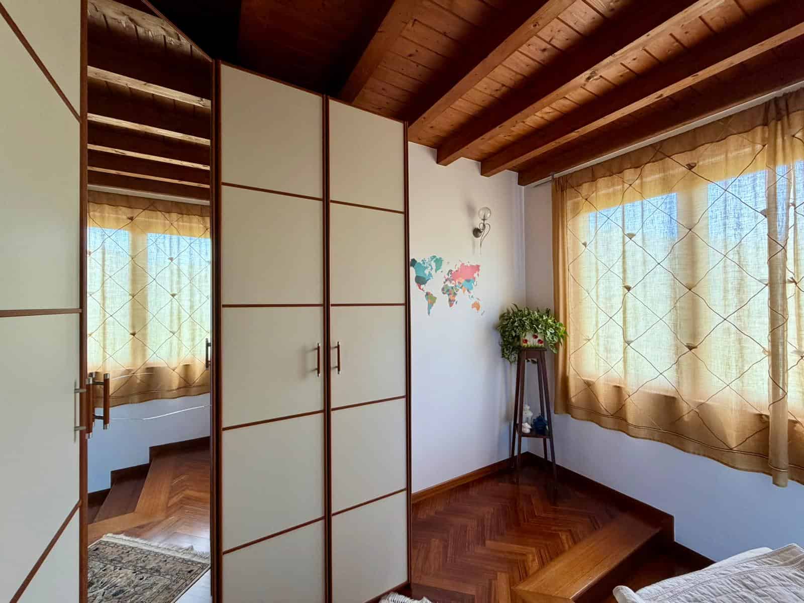 Interno di una camera con soffitto in legno, armadio a muro e grandi finestre con tende beige, vista luminosa e accogliente.