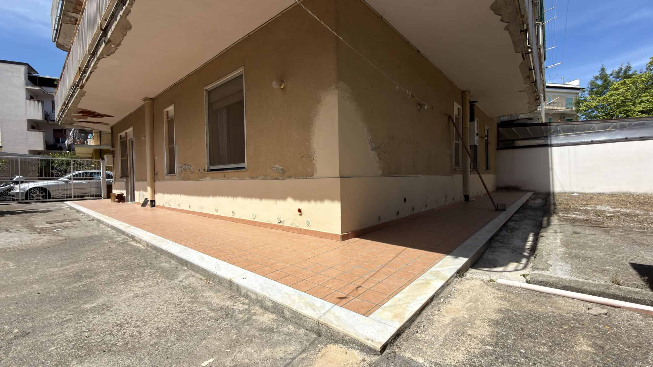 Esterno di un edificio residenziale con pareti beige, finestra e area pavimentata in piastrelle, in un contesto urbano.