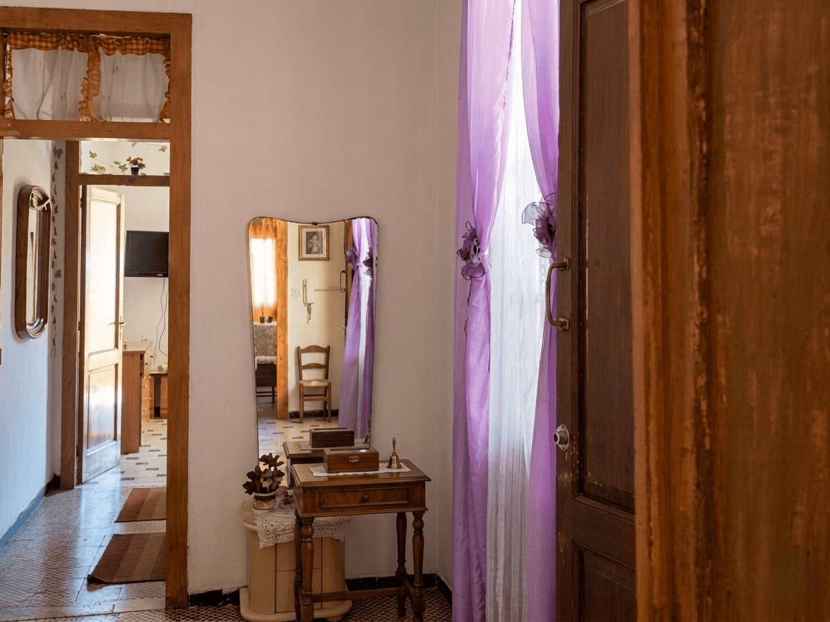 Interno di una casa con mobili in legno, specchio e tende viola, ambiente accogliente e luminoso.