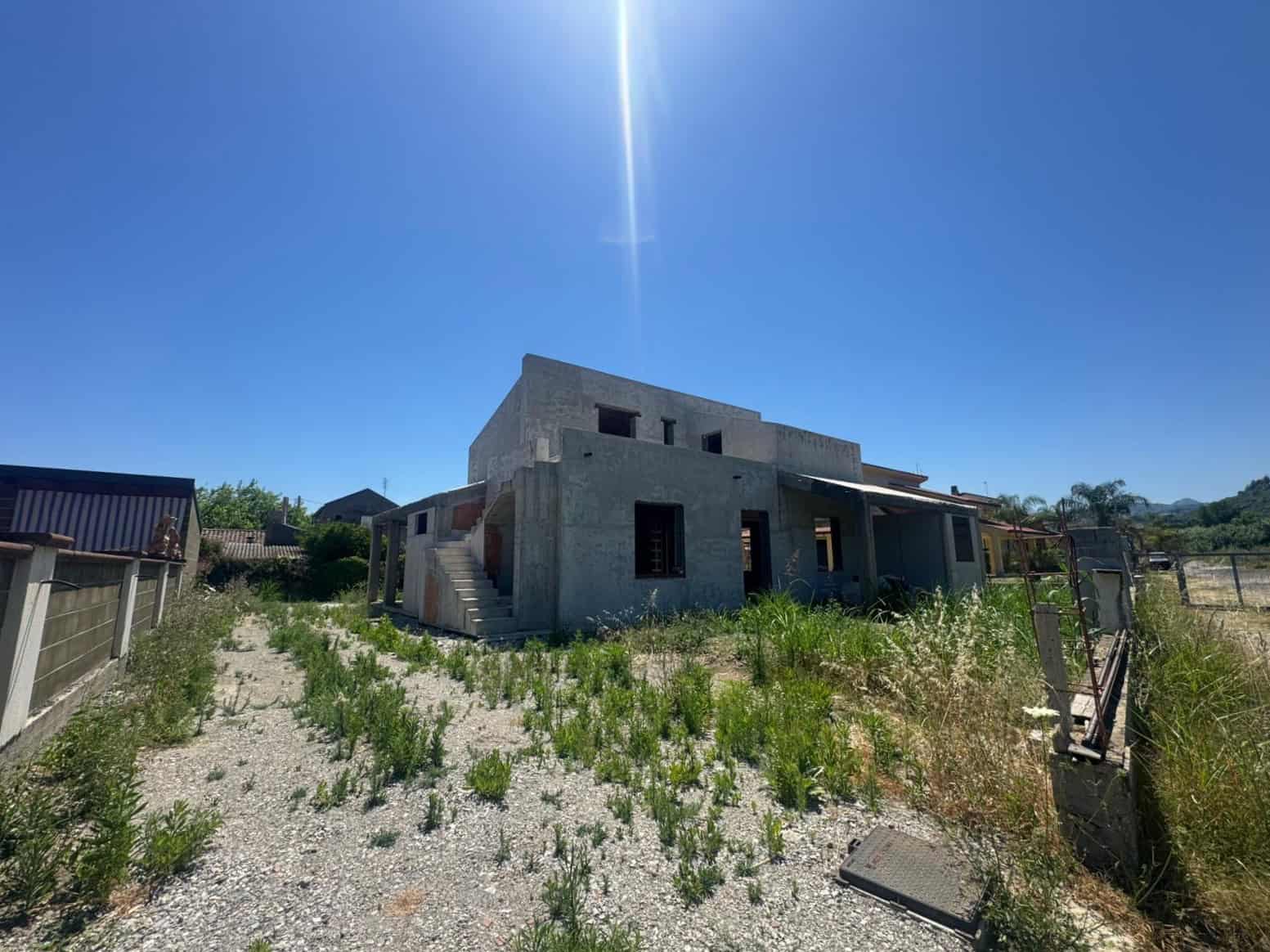 Edificio in costruzione con struttura in cemento e terreno circostante con erba e piante selvatiche sotto un cielo sereno.