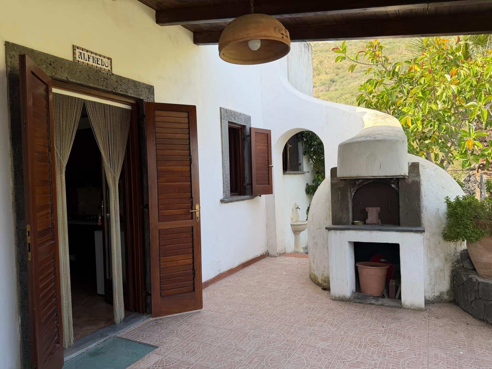 Porte in legno aperte su un'area esterna con forno a legna in stile rustico, decorata con piante verdi e un soffitto in legno, in un ambiente accogliente e tradizionale.