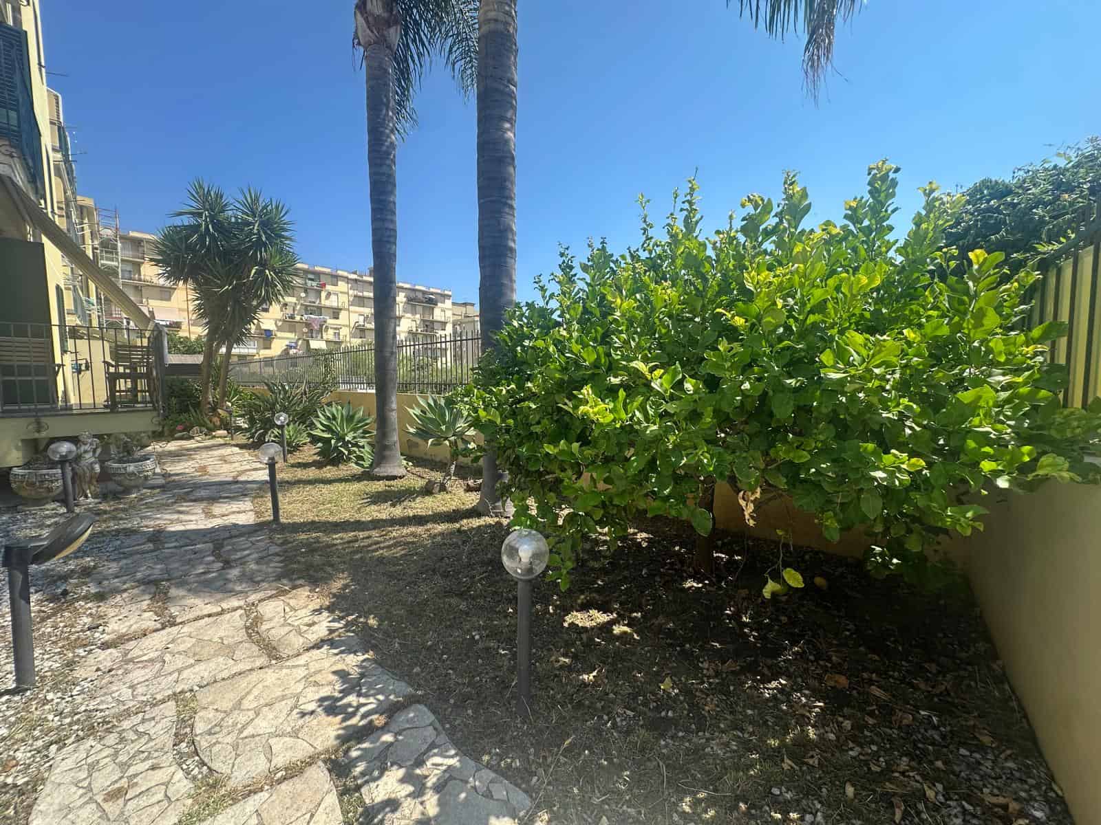 Giardino condominiale con piante di palma e arbusti, percorso pedonale in pietra, in zona residenziale di Milazzo.
