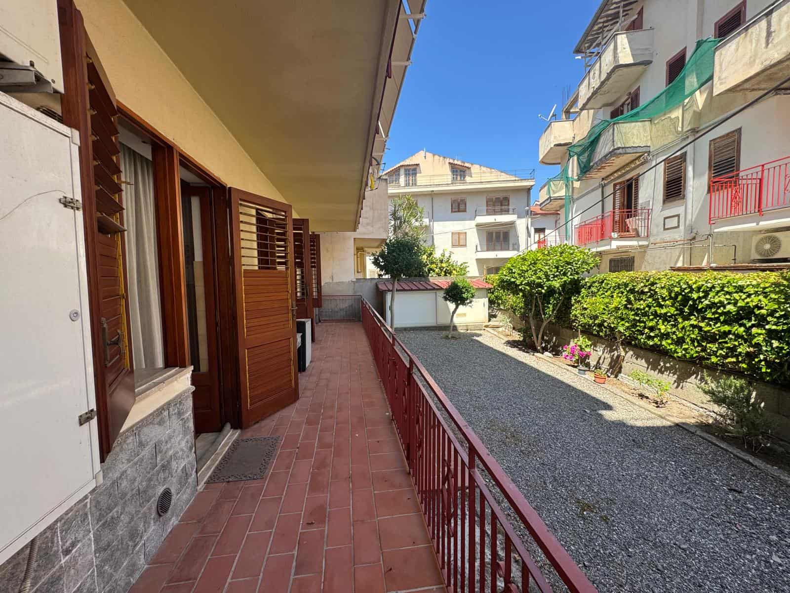 Balcone di un appartamento con ringhiera rossa, pavimentazione in piastrelle rosse, con vista su giardini e edifici residenziali in un quartiere urbano soleggiato.