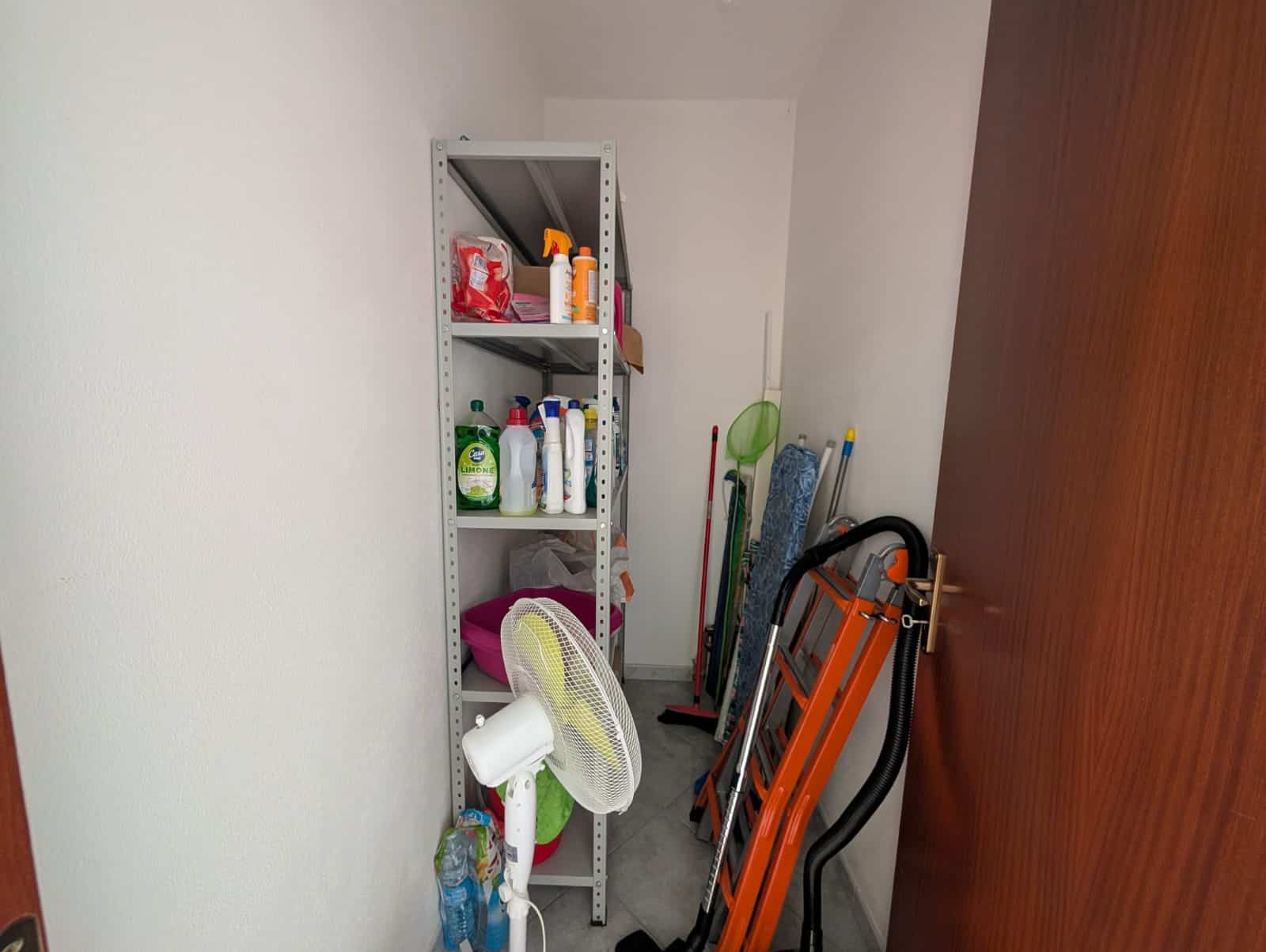 Spazio di deposito con scaffale e attrezzi per la casa, tra cui prodotti di pulizia, scope e un ventilatore, in un ambiente ordinato e funzionale.