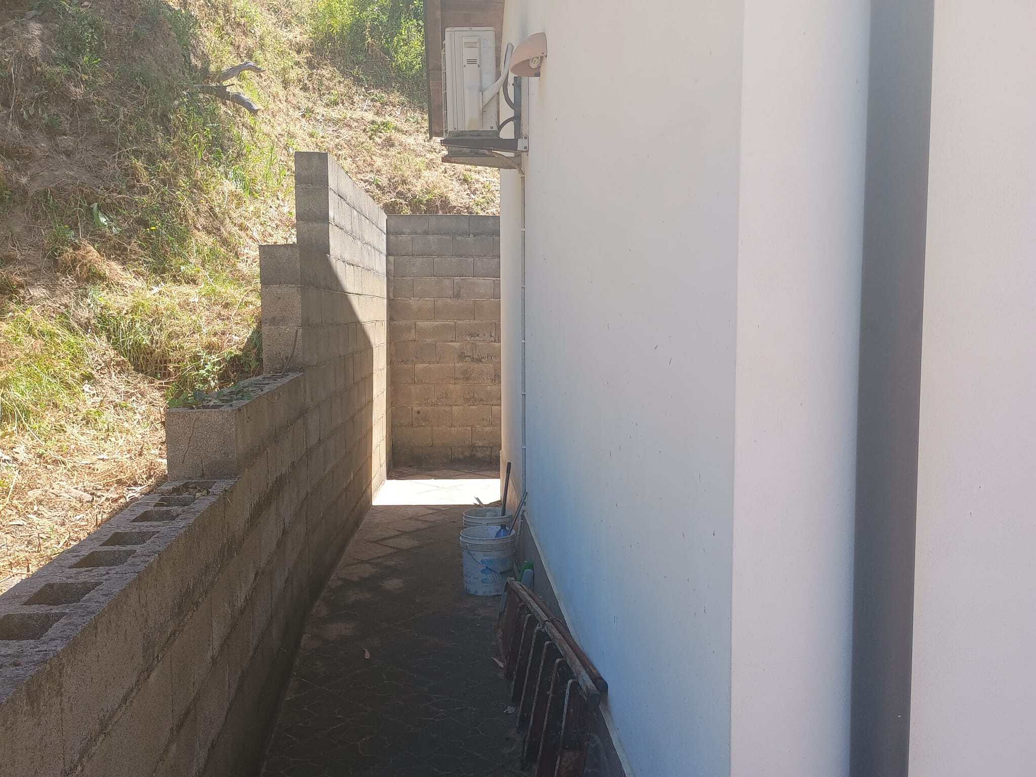 Area esterna con muro di mattoni e parete bianca, spazio di passaggio tra edificio e terreno in pendenza, con alcuni attrezzi e materiali di costruzione.
