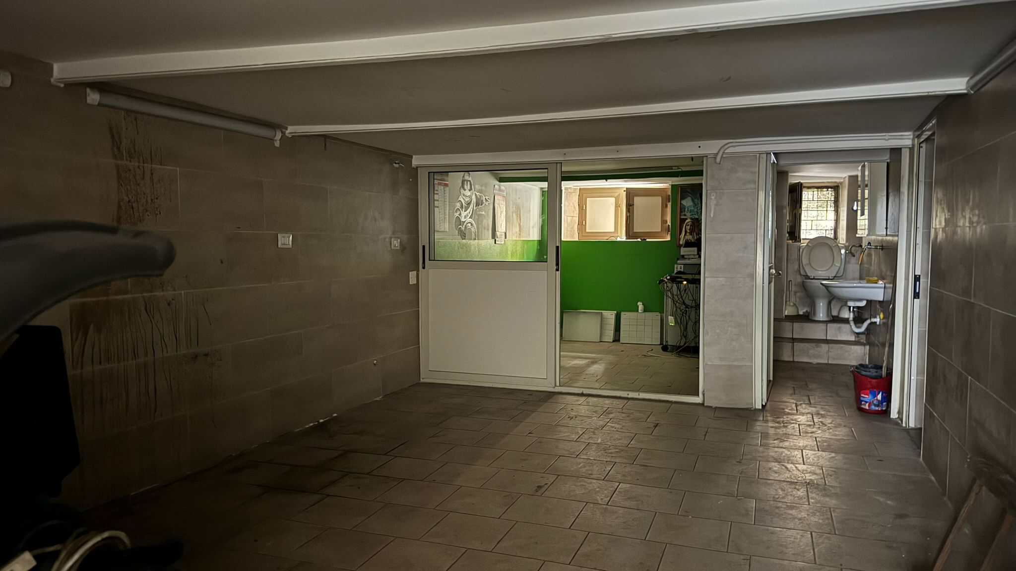 Spazio interrato con pareti rivestite in piastrelle, accesso a cucina verde, area di servizio con lavandino e tubature visibili, pavimento in piastrelle scure, ambiente funzionale e pratico.