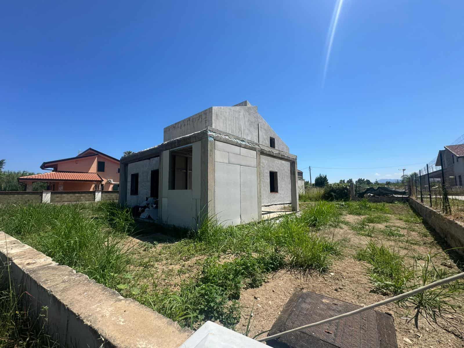 Casa in costruzione con struttura in cemento e giardino con erba verde, sotto un cielo azzurro e soleggiato.