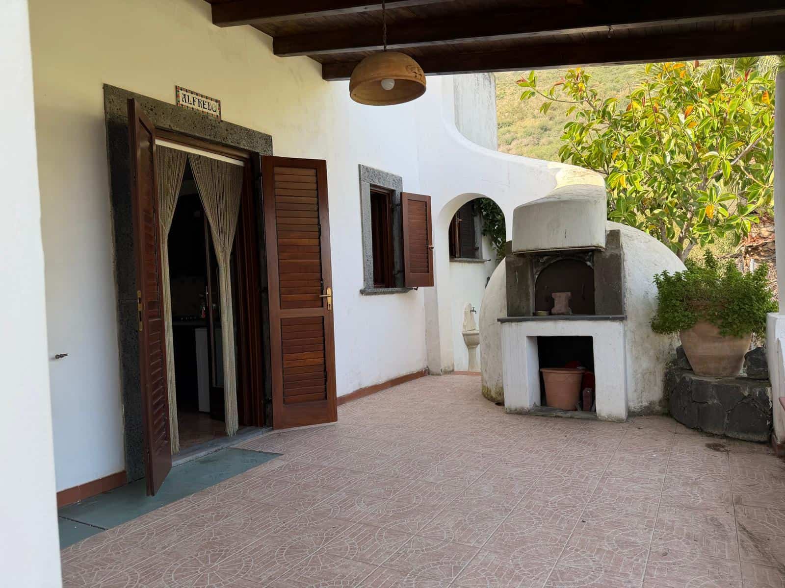 Spazio esterno di una casa mediterranea con forno a legna, pareti bianche e piante verdi, ideale per relax e cene all'aperto in stile rustico e accogliente.