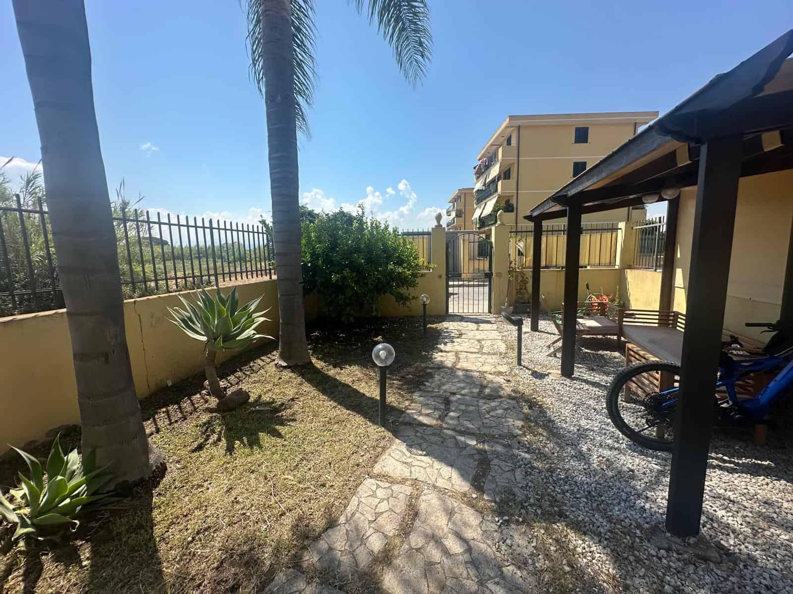 Giardino esterno di un appartamento a Milazzo con piante, gazebo e percorso in pietra, vicino a edifici residenziali.