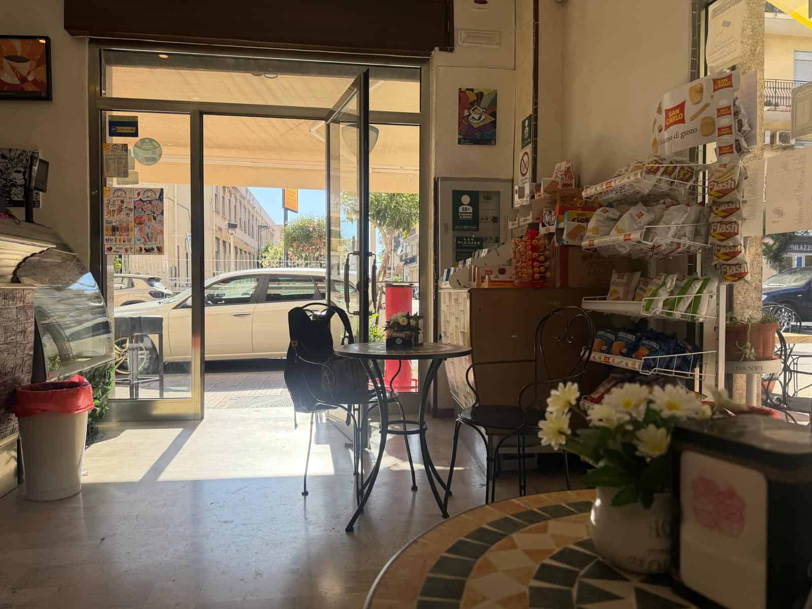 Interno di un negozio con ingresso vetrato, tavoli e scaffali con prodotti alimentari e snack, atmosfera luminosa e accogliente.