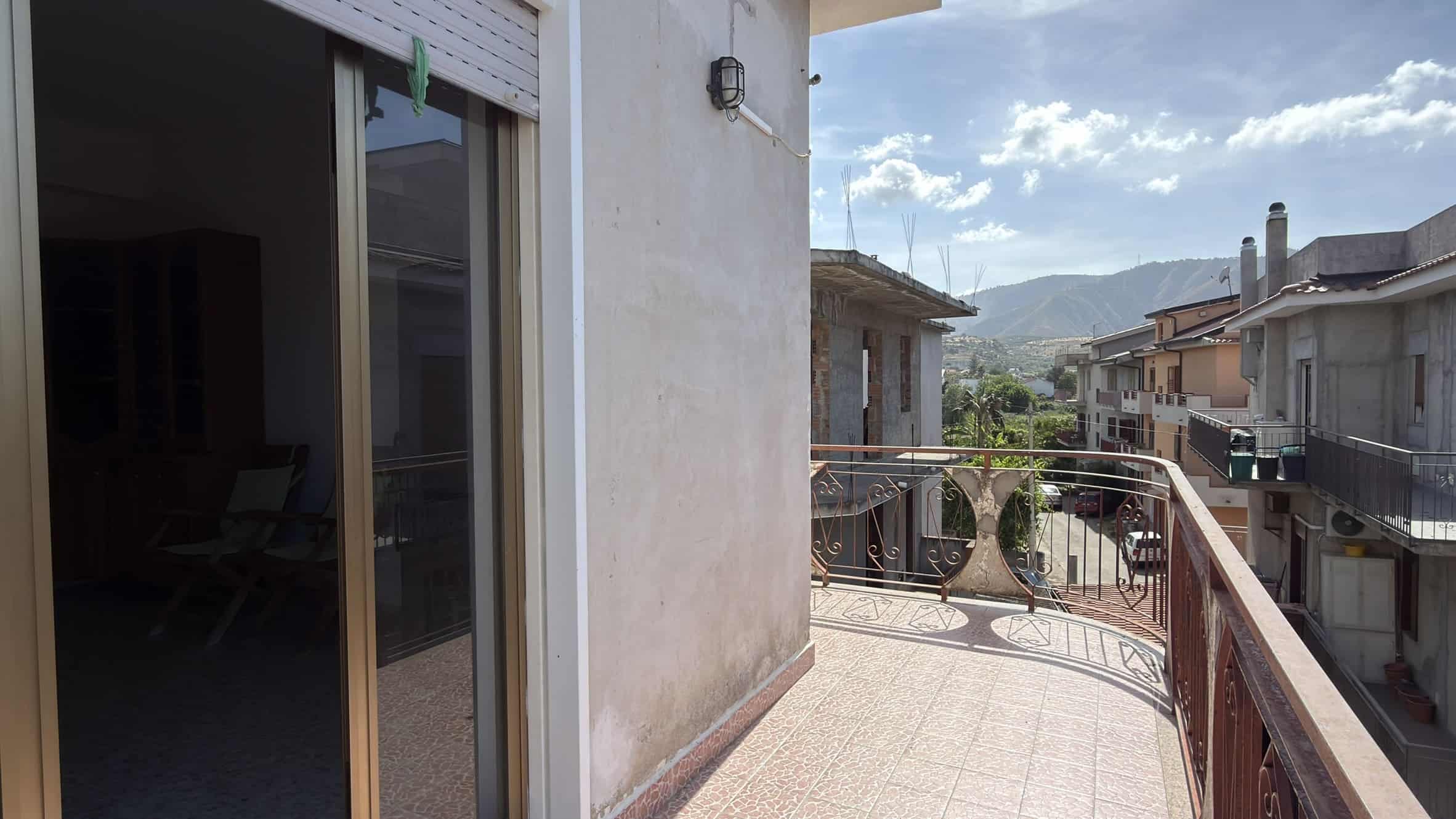 Balcone di un appartamento con vista sulle case e sulle montagne sullo sfondo, in una giornata soleggiata.