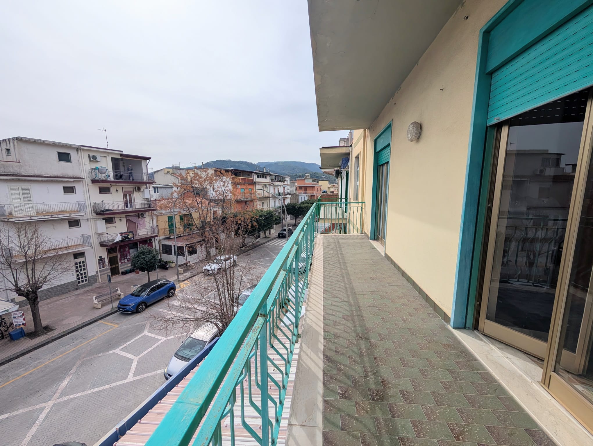 Balcone di un appartamento con vista sulla strada e edifici circostanti, in un giorno nuvoloso.