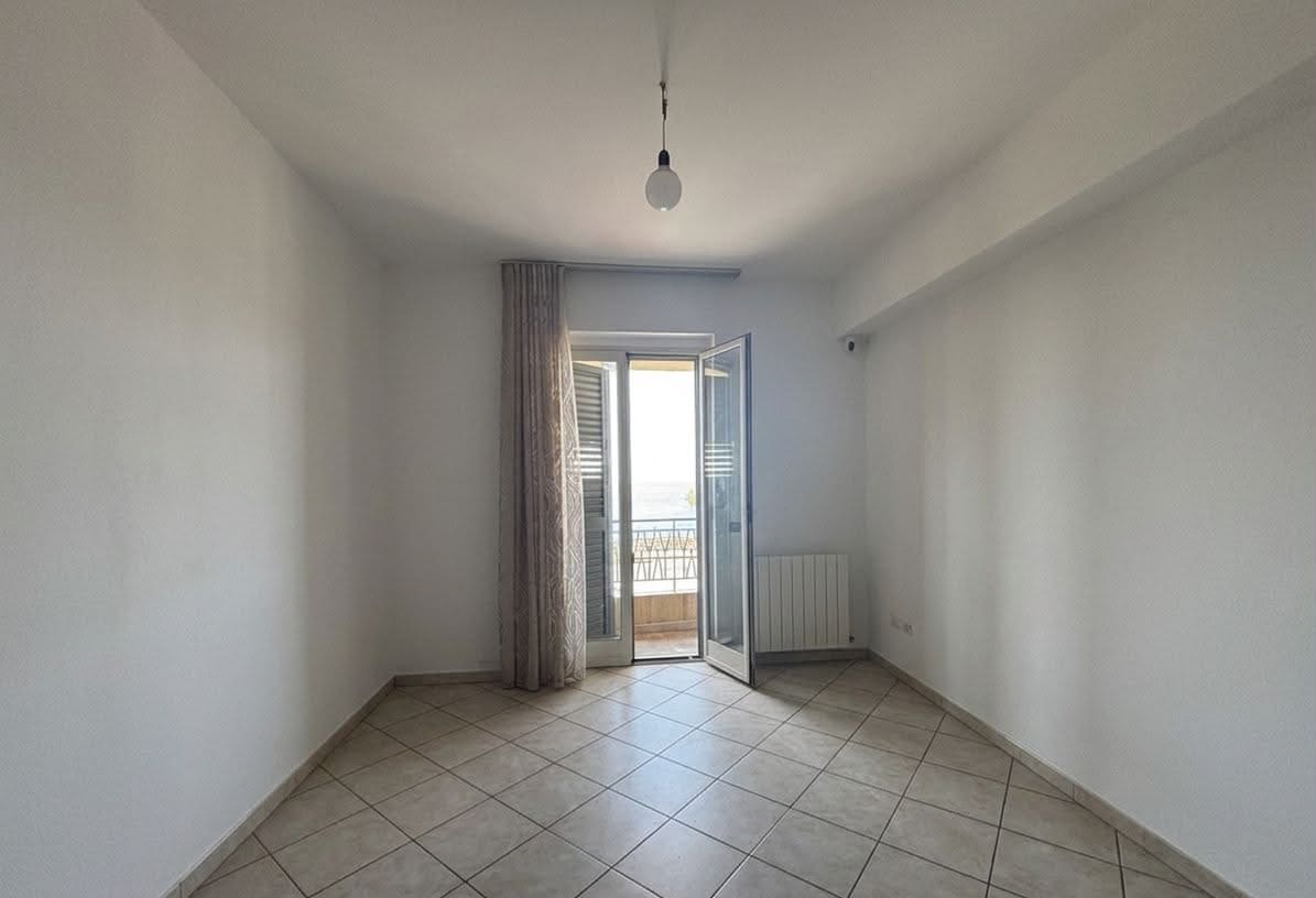 Stanza vuota con porta finestra che apre su un balcone con vista mare, pavimento in piastrelle e pareti bianche.