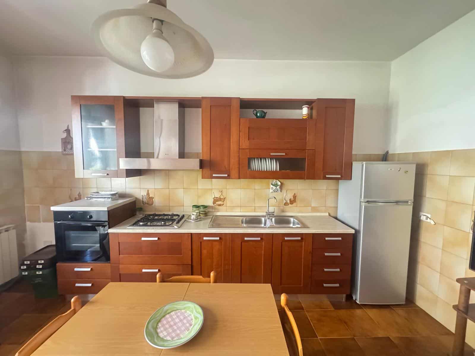 Cucina compatta con mobili in legno, forno, frigorifero e tavolo con sedie in un ambiente domestico accogliente.