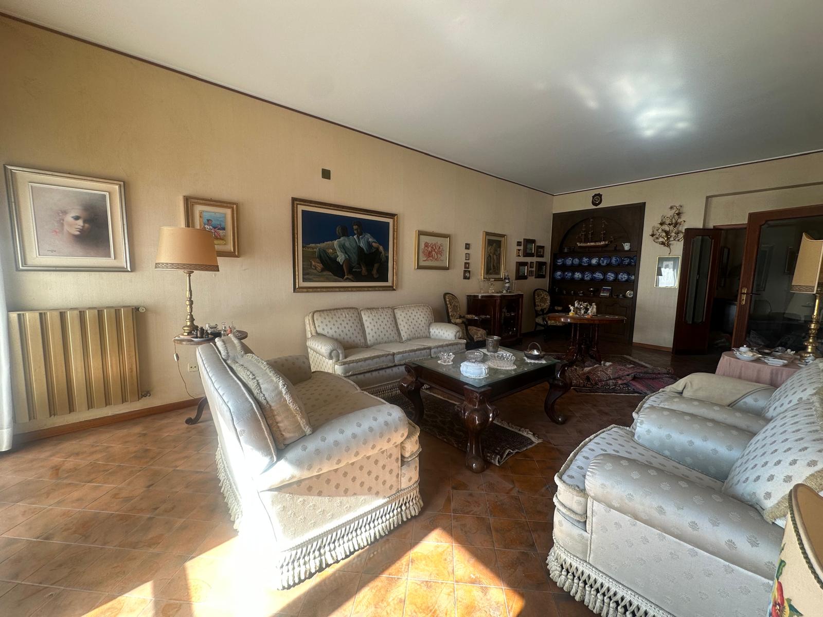 Elegante soggiorno con divani in tessuto chiaro, tavolino in legno, lampada da terra e pareti decorate con quadri, ambiente accogliente e raffinato, ideale per momenti di relax e convivialità.