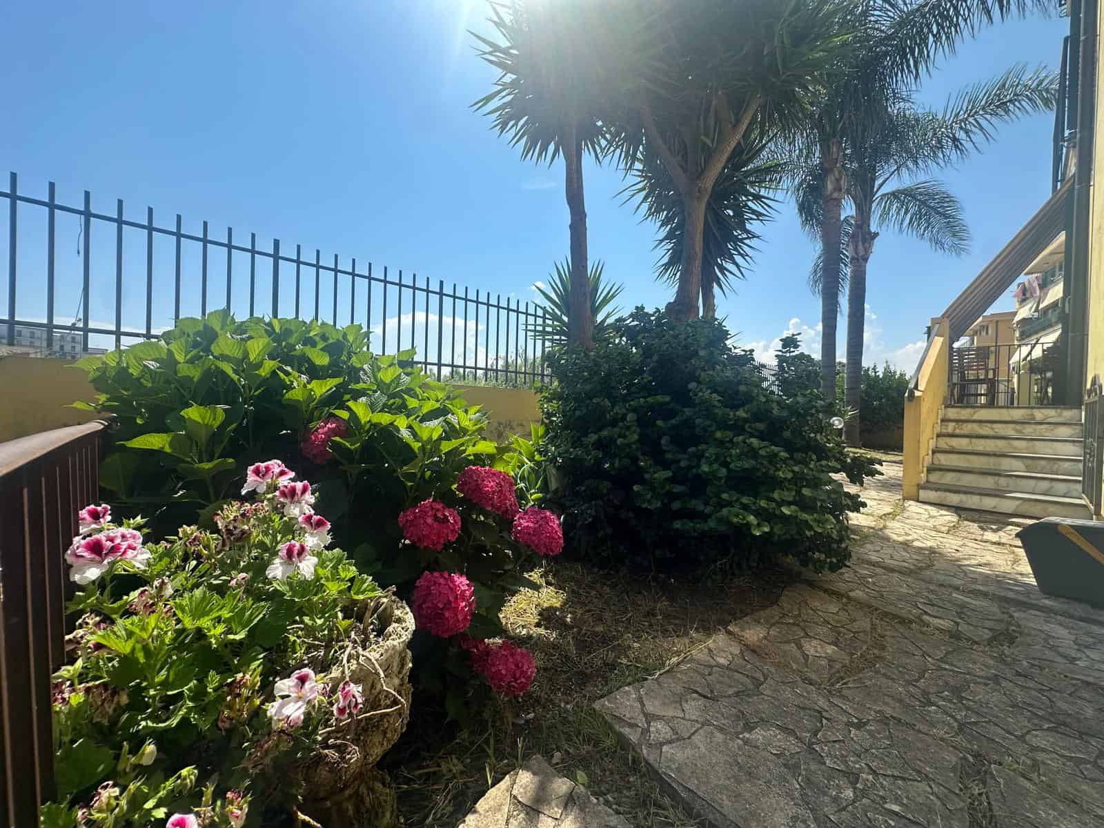 Giardino con palme e fiori colorati in un appartamento in vendita a Milazzo, vista soleggiata e tranquilla.