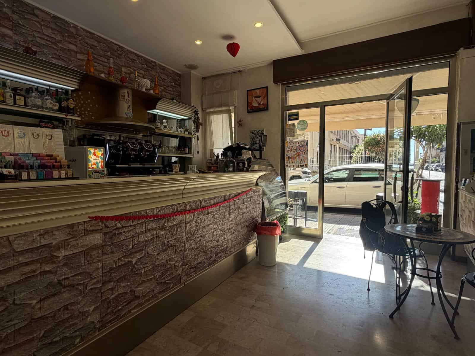 Interno di un bar con bancone in pietra e area con tavoli e sedie, luce naturale dall'ingresso.