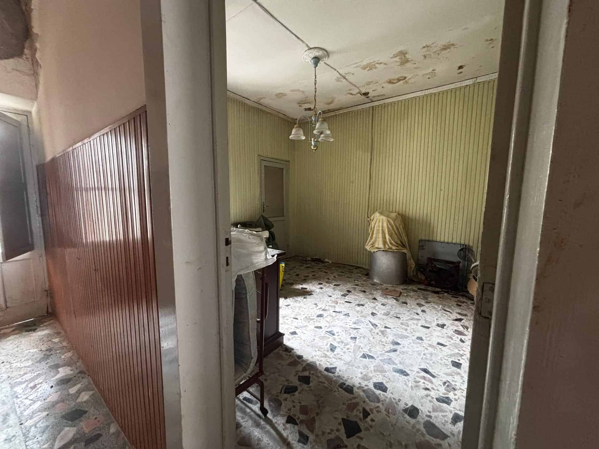 Interno di una stanza in condizioni di abbandono con pareti a strisce verdi, soffitto danneggiato e pavimento in piastrelle di pietra, con mobili e oggetti abbandonati.