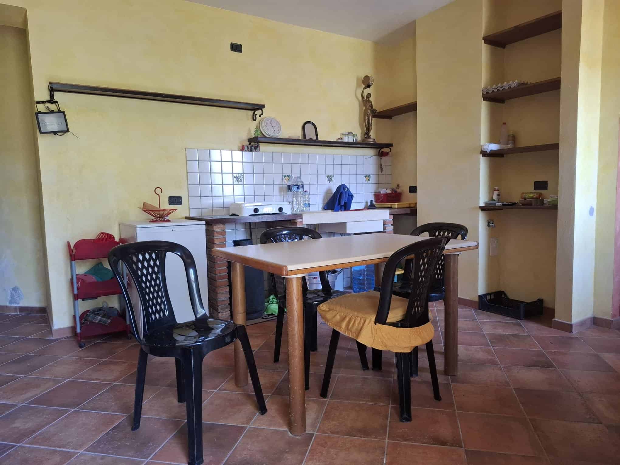 Una cucina accogliente con tavolo in legno, sedie nere e scaffali aperti, in un ambiente luminoso con pareti gialle e pavimento in piastrelle di ceramica.
