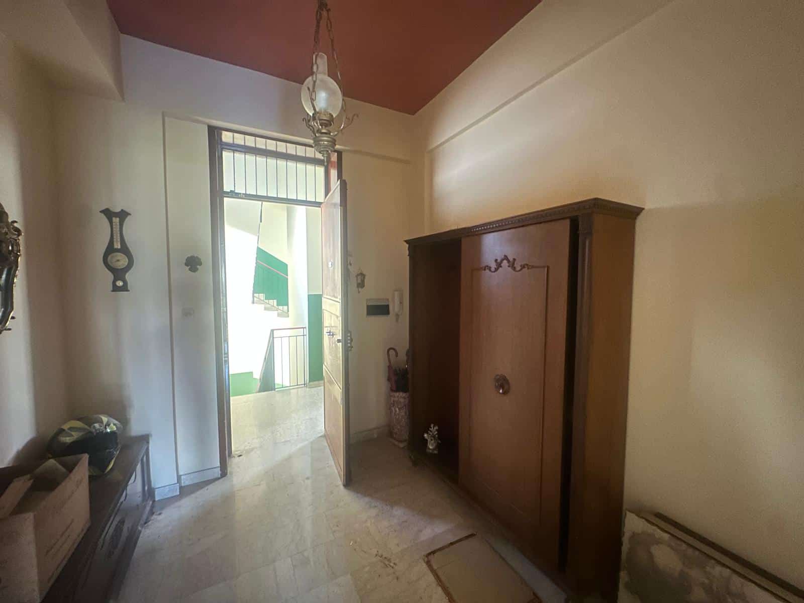 Armadio in legno antico in un ingresso di casa con pareti beige e soffitto rosso, illuminato da luce naturale e lampadario vintage.
