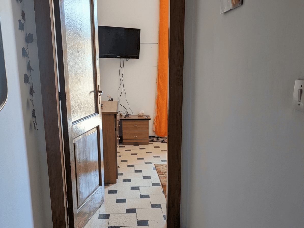 Un ingresso di casa con porta in legno, vista su soggiorno con TV, pavimento in piastrelle bianche e nere, pareti chiare e decorazioni colorate, ambiente accogliente e luminoso.