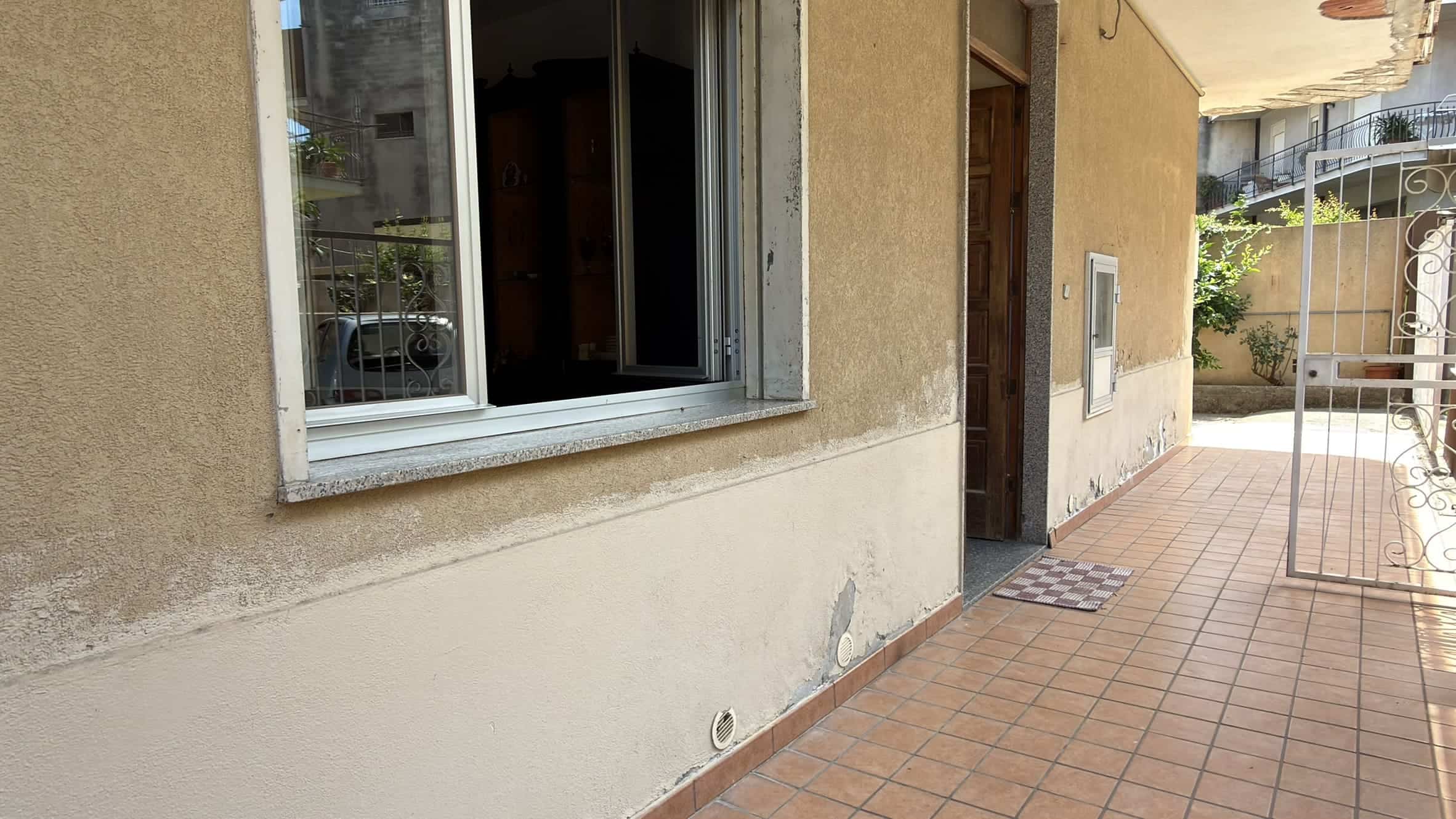 Esterno di una casa con pareti beige, finestra e porta d'ingresso, pavimentazione in piastrelle e cancello in ferro battuto.