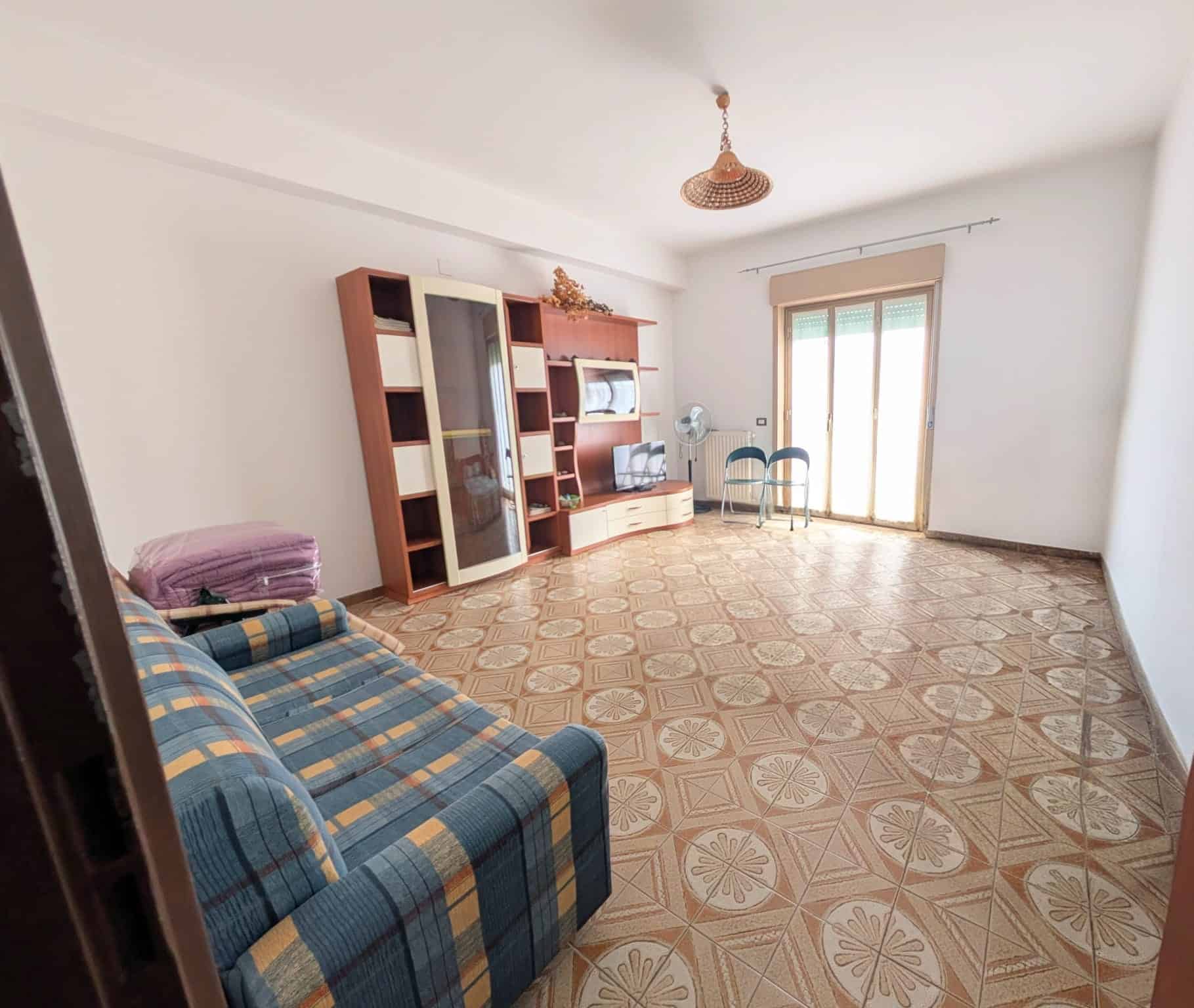 Soggiorno spazioso con divano a righe, libreria e accessori, luce naturale da porta finestra, pavimento in piastrelle decorato, arredamento semplice.