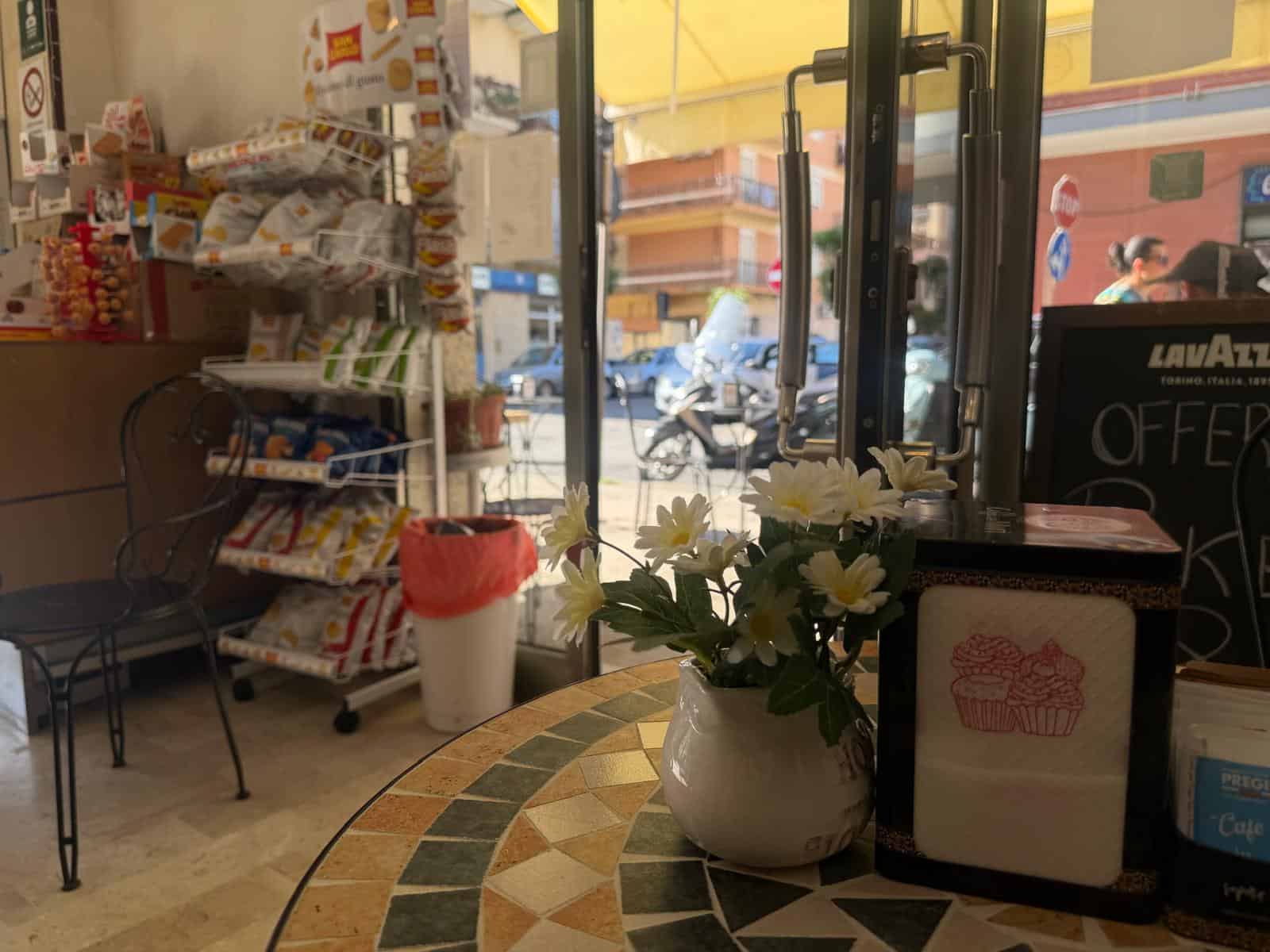 Interno di un negozio con vetrina, fiori in vaso e scaffali con prodotti alimentari.