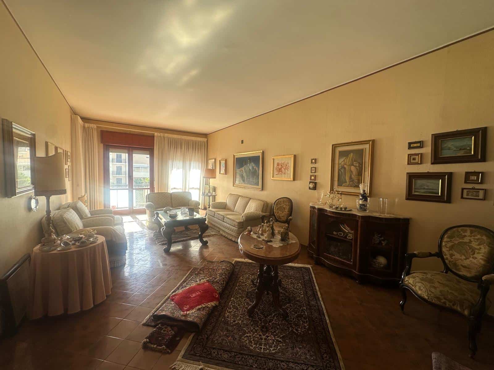 Elegante soggiorno con mobili classici, quadri alle pareti e grande finestra con tende leggere, arredato con divani, poltrone e tavolini in stile tradizionale, ideale per vivere momenti di relax e con.