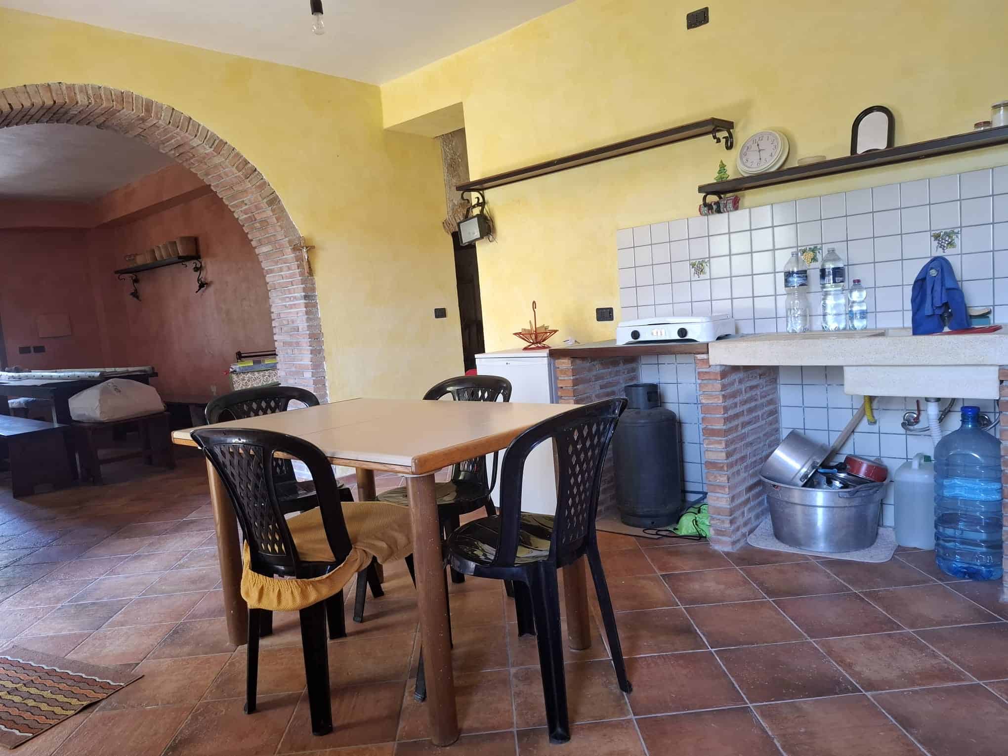 Un angolo cucina in stile rustico con tavolo in legno, sedie nere e pareti gialle, dotato di lavandino, scaffali e vari contenitori, ideale per ambienti abitativi accoglienti e funzionali.