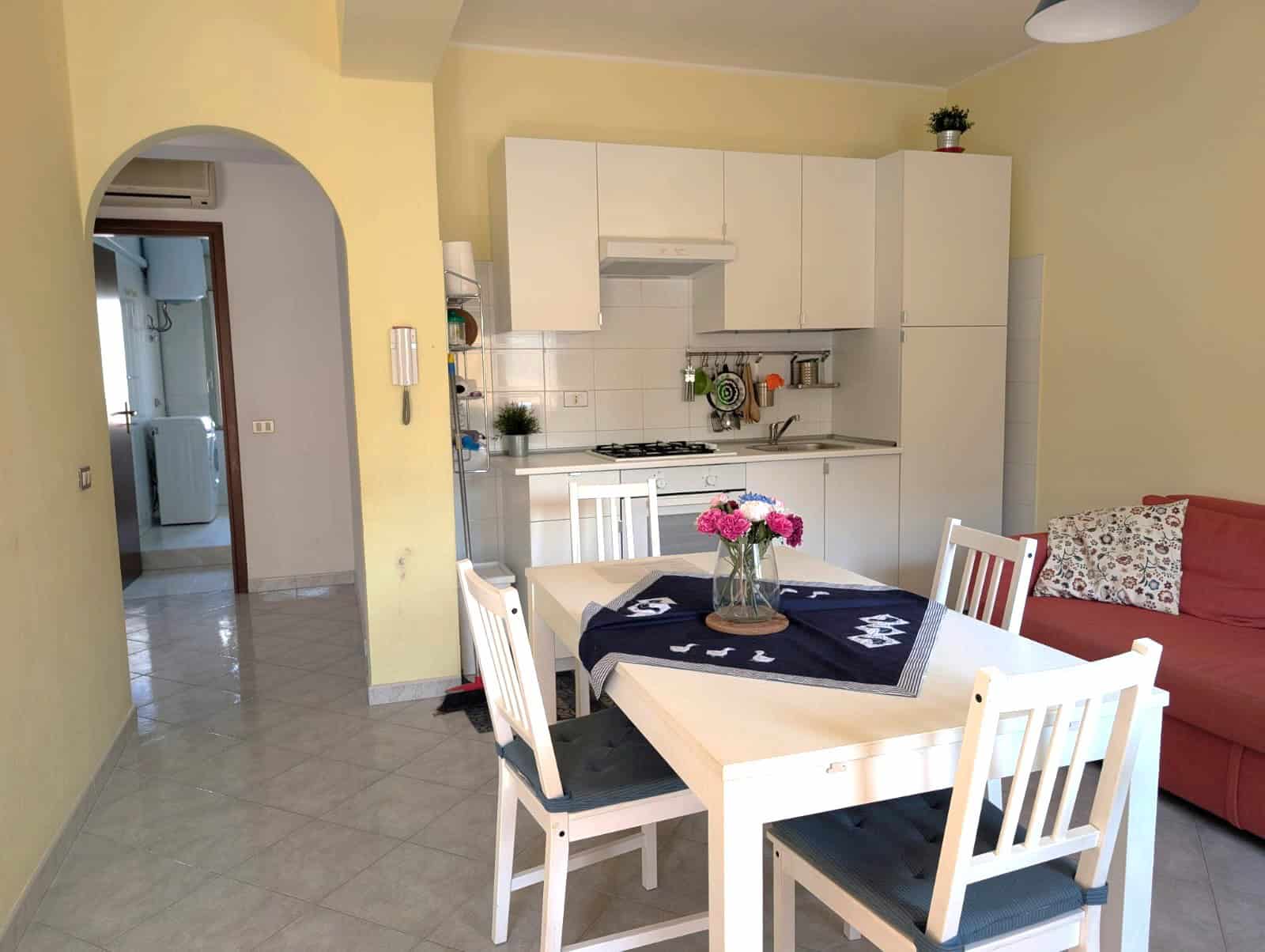 Interno di un appartamento con cucina moderna, zona pranzo con tavolo e sedie, e soggiorno con divano rosso, ambiente luminoso e accogliente.