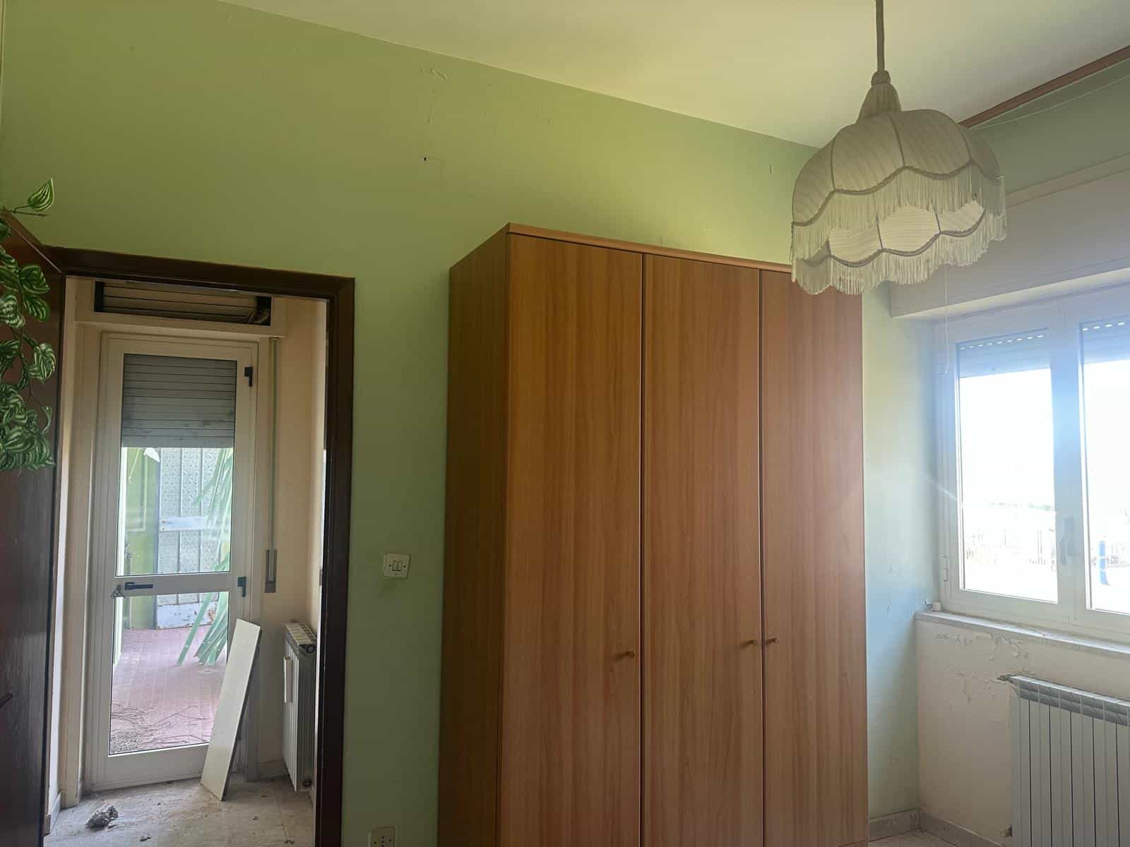 Armadio in legno in una stanza luminosa con pareti verde chiaro, finestra e porta finestra che conduce a un balcone, illuminata da lampadario a soffitto con lampada a forma di nuvola.