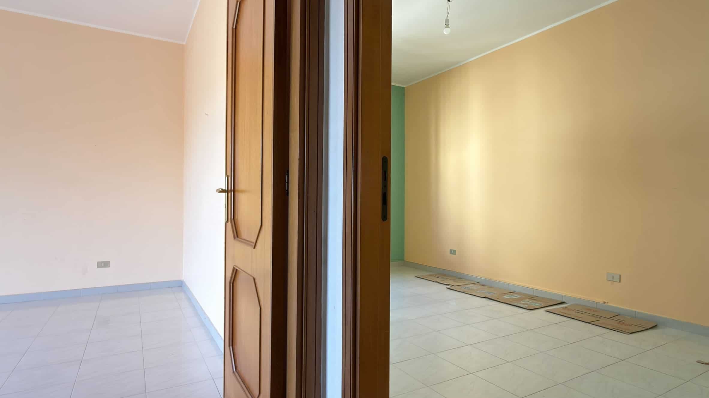 Interno di un appartamento con pareti beige e pavimento in piastrelle chiare, vista da una porta aperta verso un soggiorno vuoto.