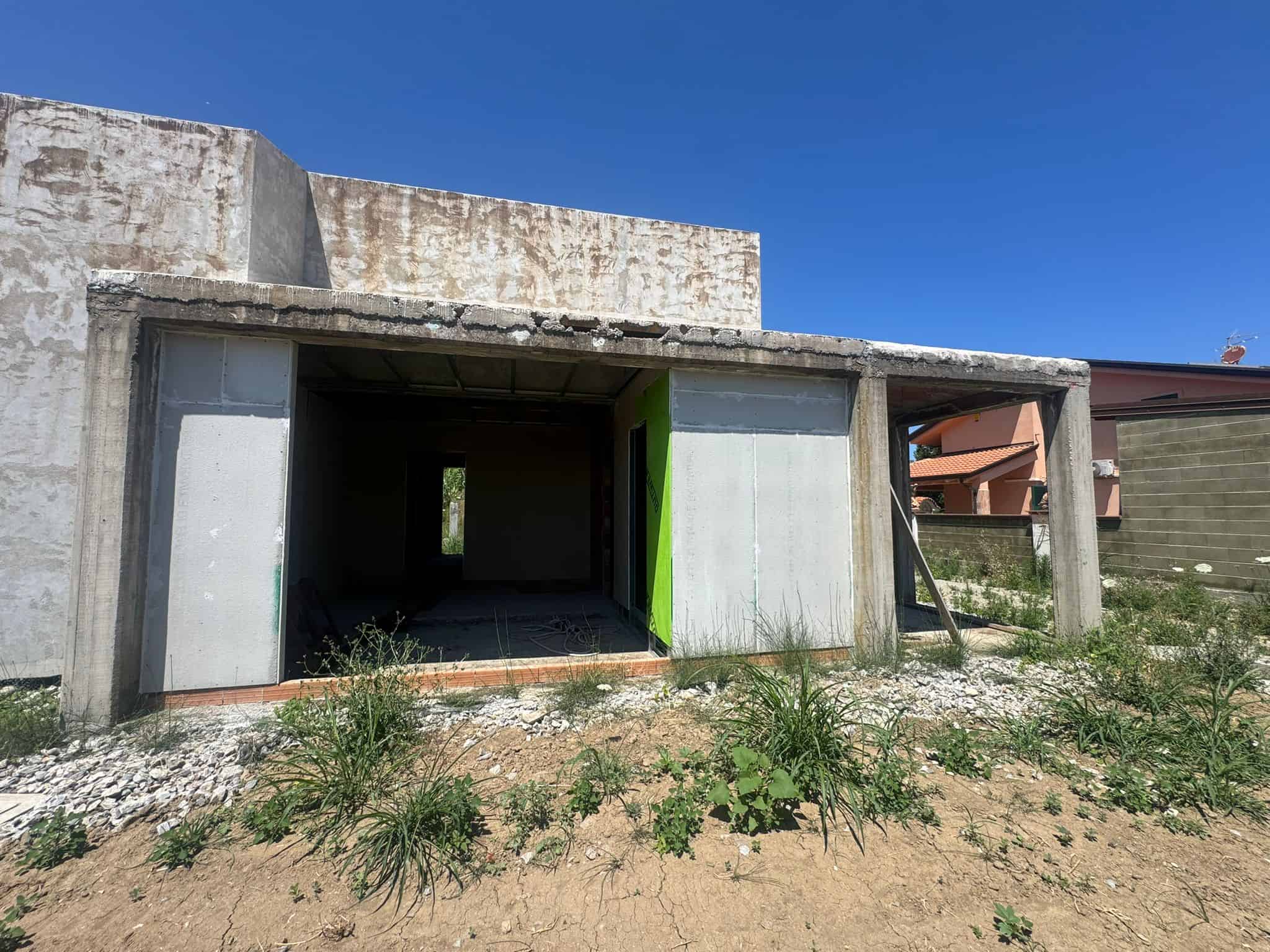 Struttura di casa in costruzione con pareti in cemento e apertura per porta e finestre, su terreno con vegetazione e cielo blu.