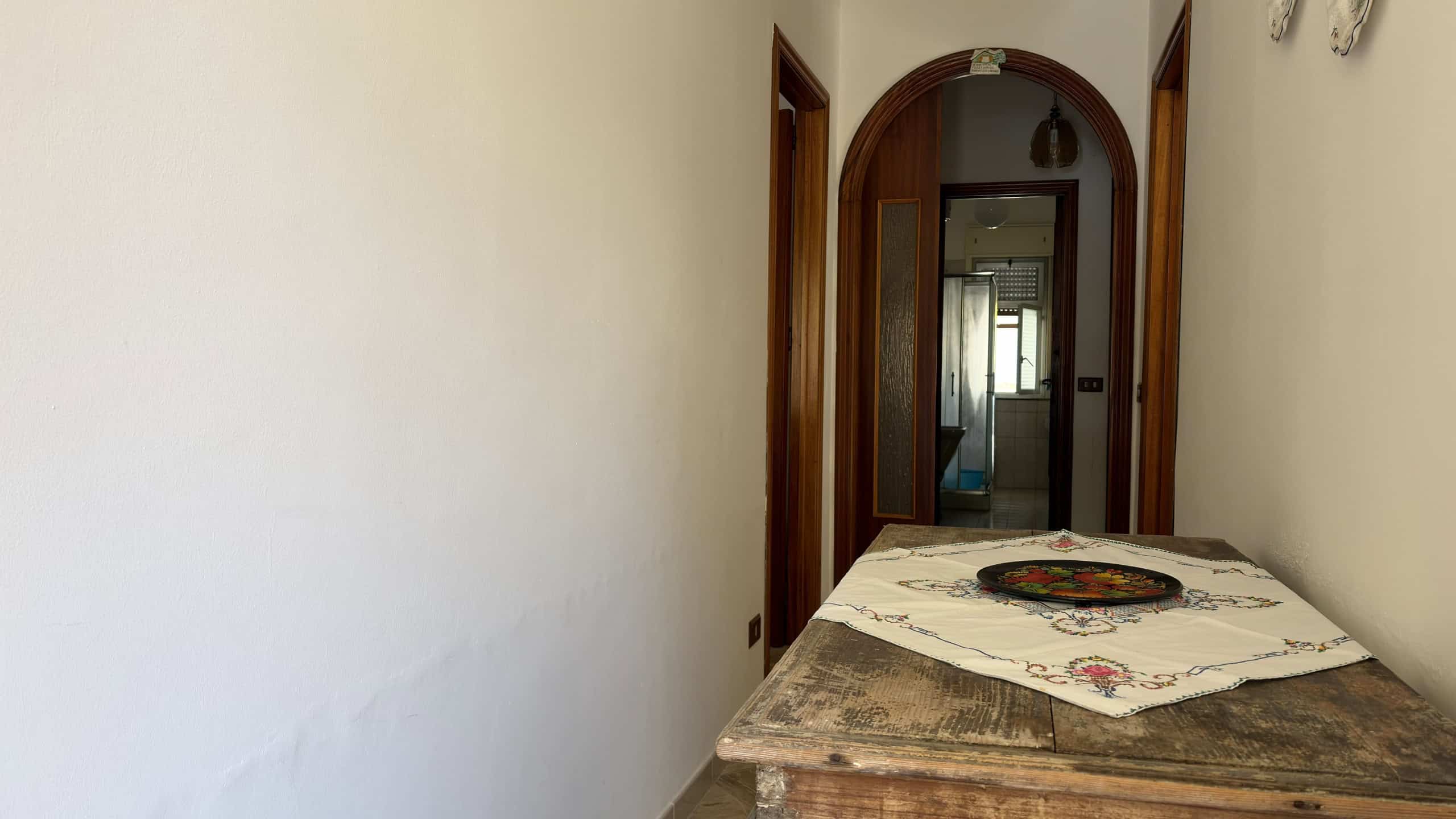 Interno di un appartamento con porta d'ingresso in legno e tavolo rustico con tovaglia decorativa, ambiente luminoso e semplice.