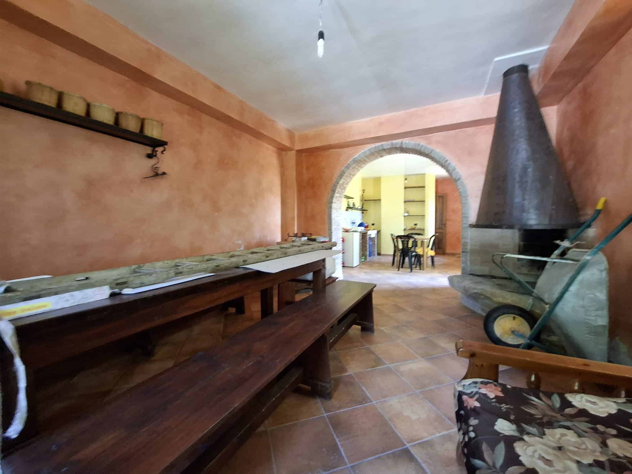 Ampia sala rustica con camino in pietra, pareti color terracotta, tavolo in legno e sedie, ideale per eventi o riunioni familiari, con accesso a cucina aperta e spazio accogliente.