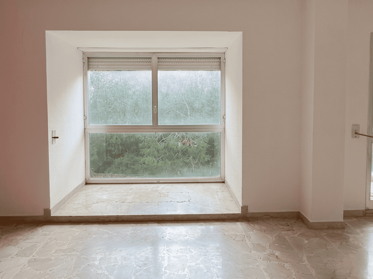 Vetrata grande con vista su giardino verde, luce naturale che entra in una stanza vuota, pavimento in marmo chiaro, pareti bianche, ambiente luminoso e spazioso, ideale per ristrutturazione o arredame.