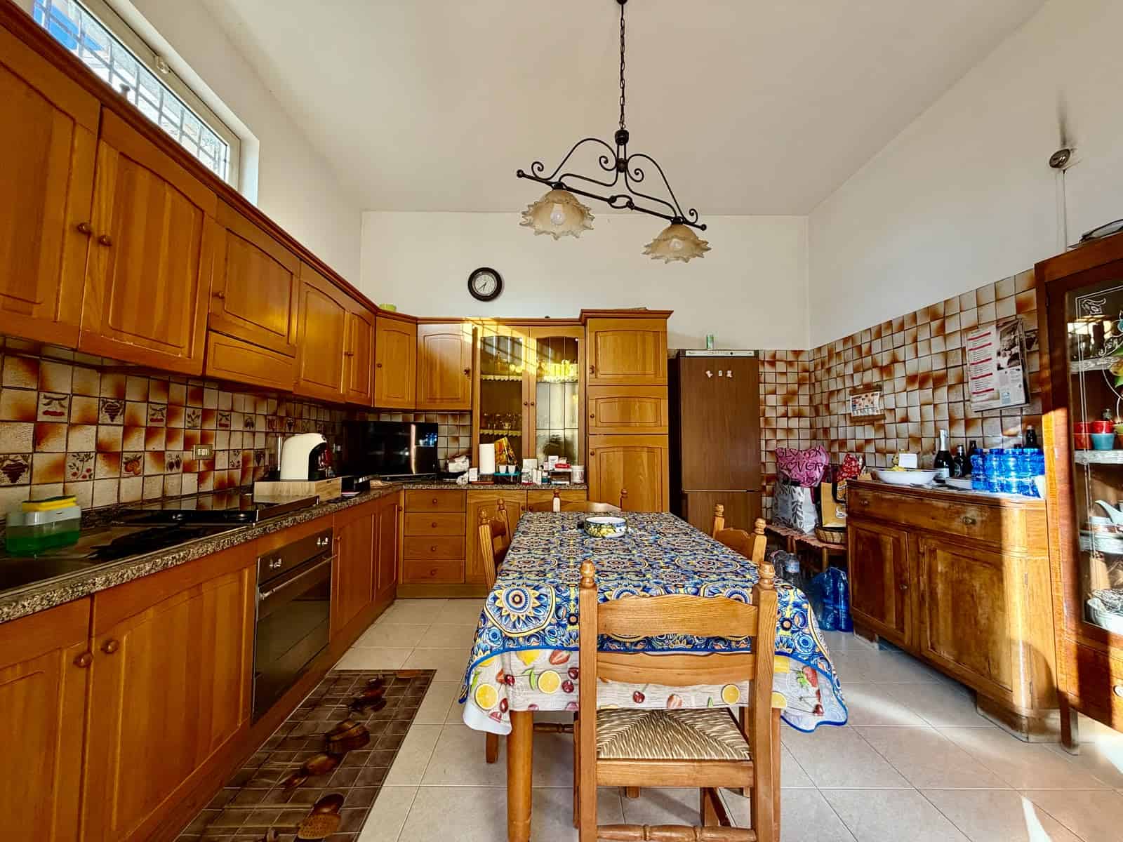Cucina spaziosa con mobili in legno, tavolo con tovaglia colorata e illuminazione centrale.