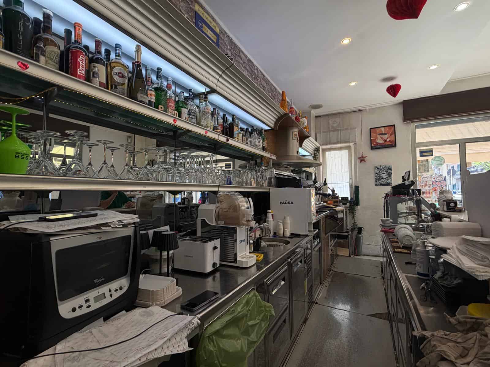 Interno di un bar con bancone, bottiglie di alcolici e attrezzature da bar, ambiente luminoso e accogliente.