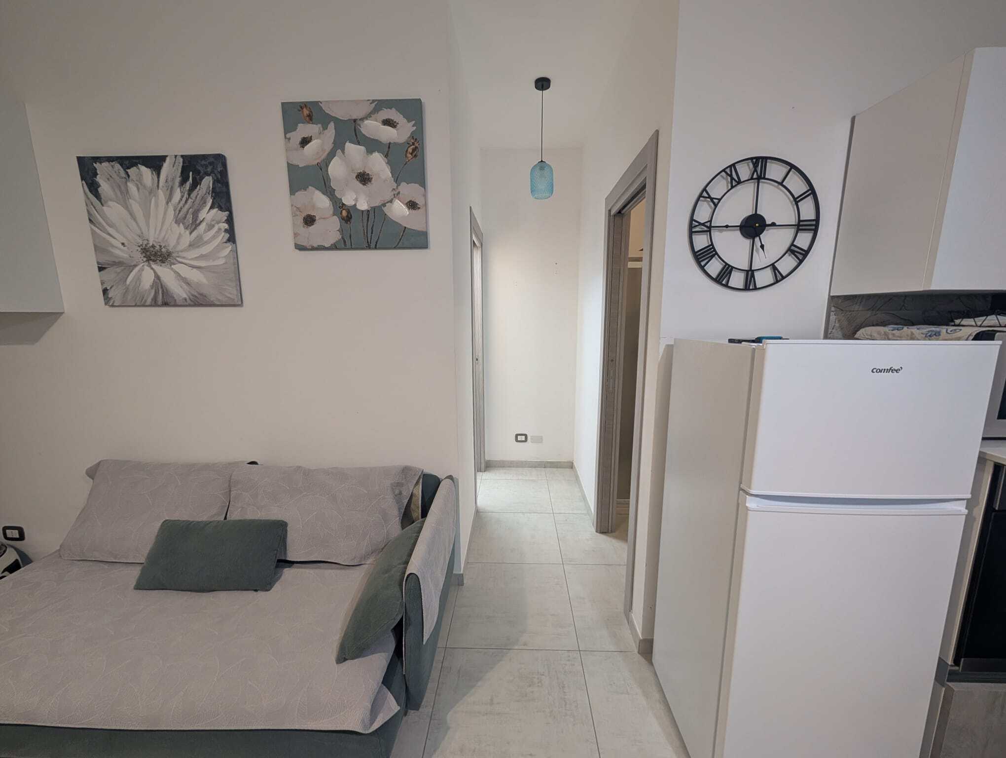 Elegante soggiorno con divano grigio, decorazioni floreali alle pareti e un grande orologio nero, in un ambiente luminoso e moderno.