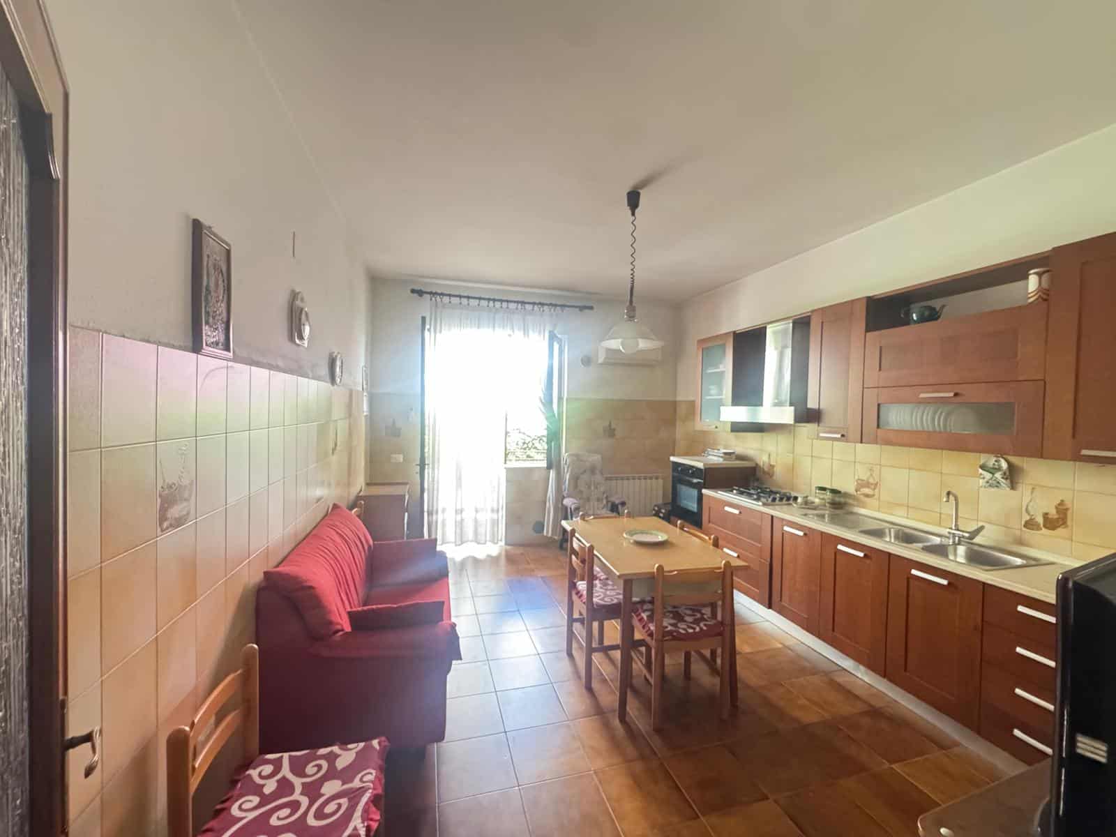 Cucina moderna con mobili in legno, tavolo da pranzo e accessori, ambiente luminoso e accogliente con porta finestra che dà su un balcone o terrazza.