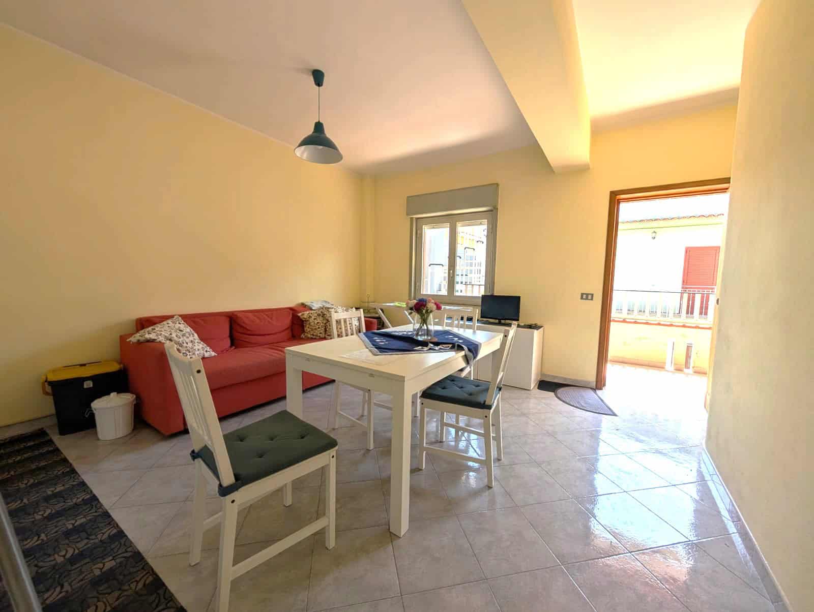 Casa con soggiorno luminoso e balcone soleggiato, arredato con divano rosso, tavolo bianco e sedie, ideale per relax e convivialità. Ampie finestre e accesso al balcone offrono luce naturale e vista s.