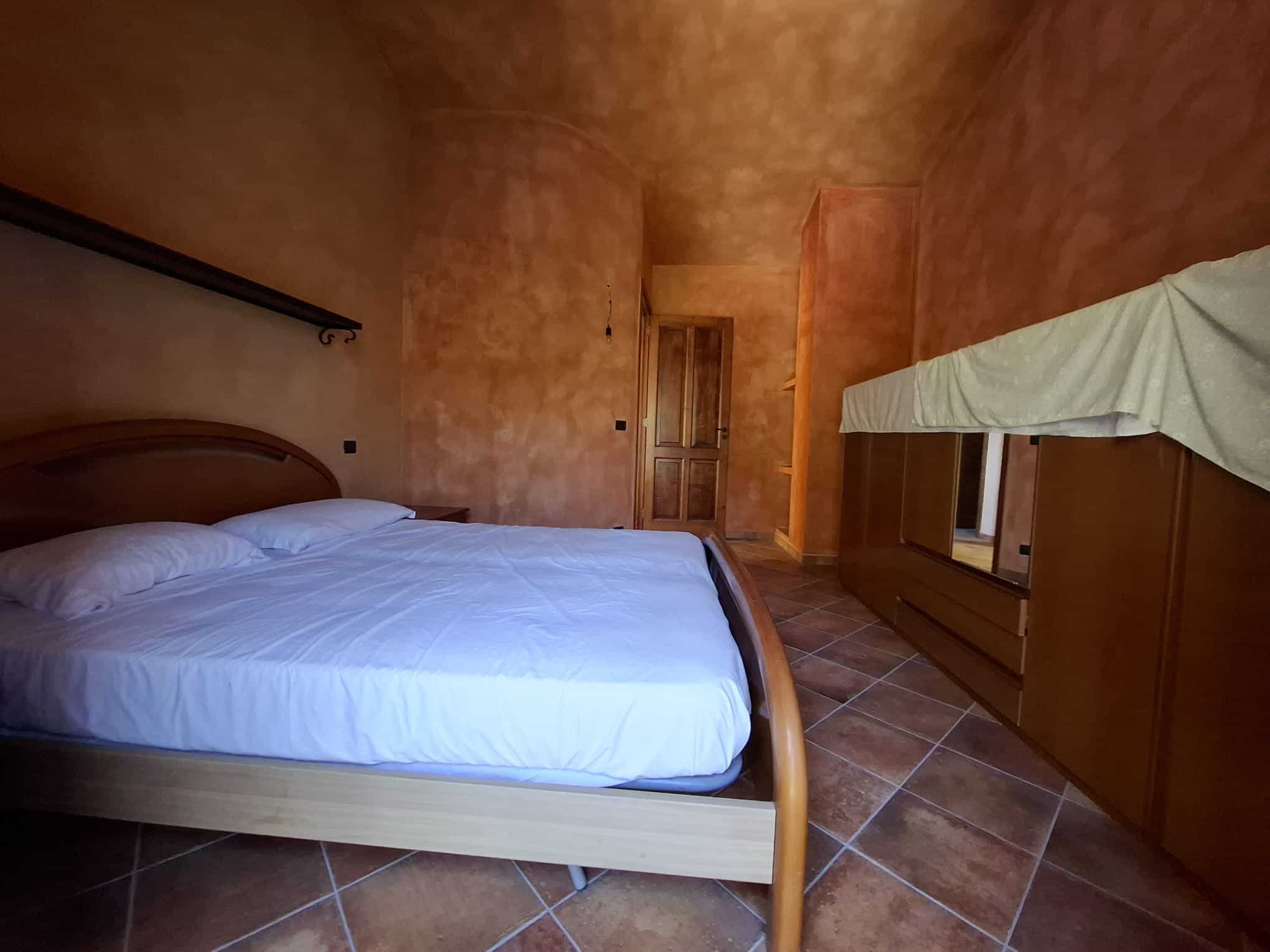 Il letto matrimoniale in una camera con pareti color terracotta, arredi in legno e pavimento in piastrelle, ambiente accogliente e rustico, ideale per soggiorni rilassanti.