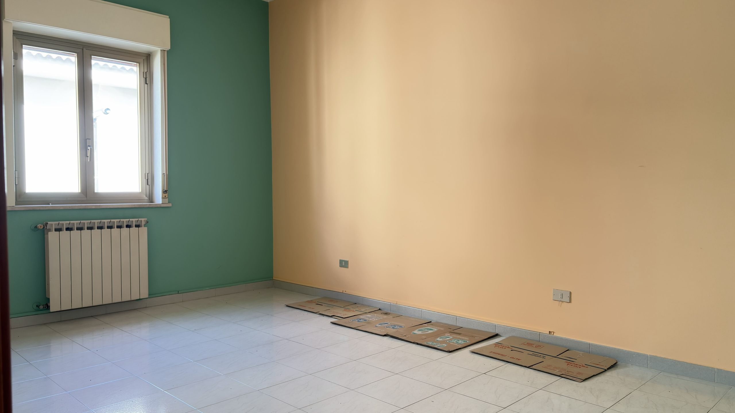 Stanza vuota in un appartamento con pareti verde e beige, finestra e radiatore, pavimento in piastrelle e alcuni cartoni sul pavimento.