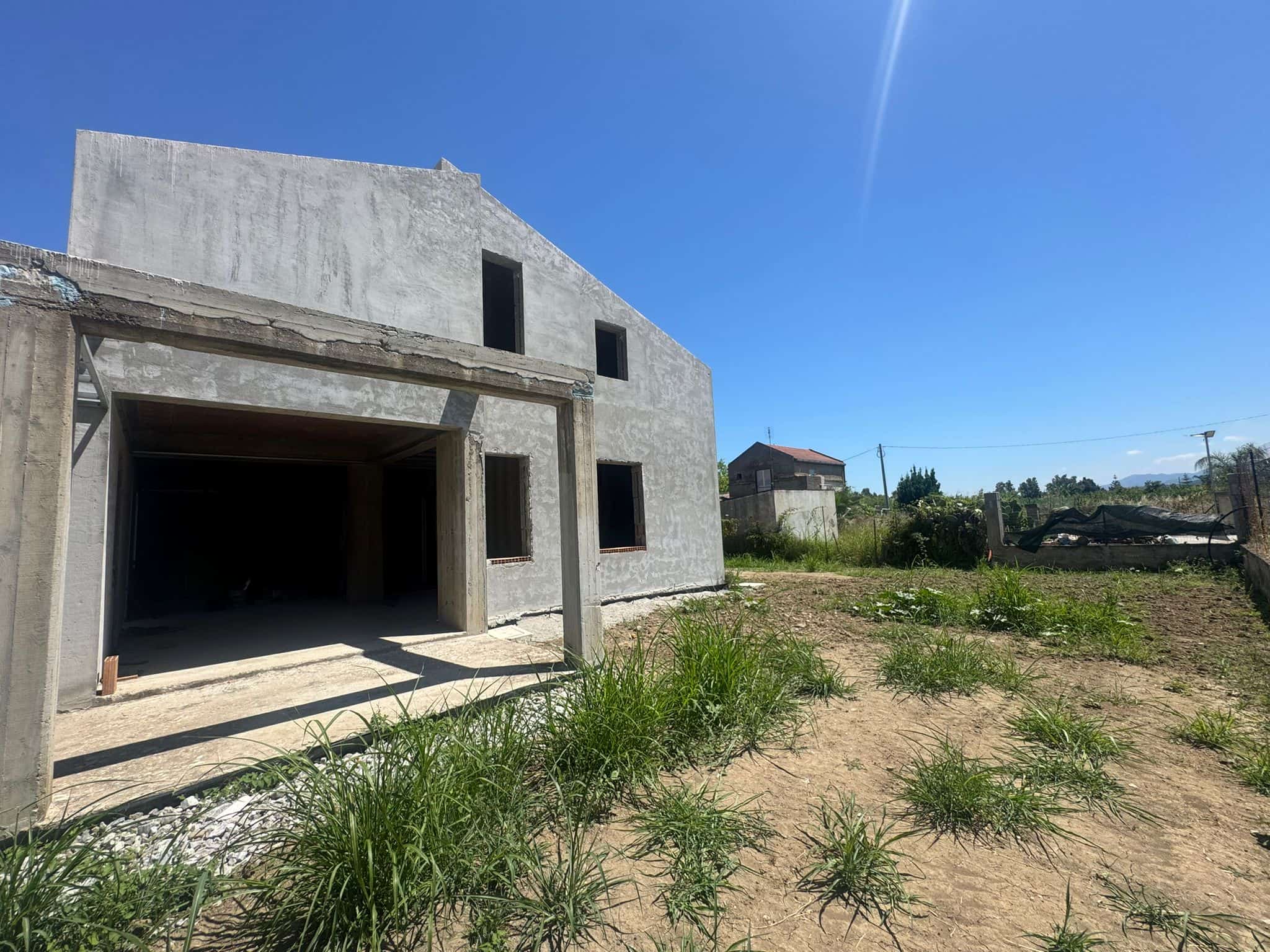 Casa in costruzione con struttura in cemento e spazio esterno con erba e terreno sabbioso sotto un cielo sereno.