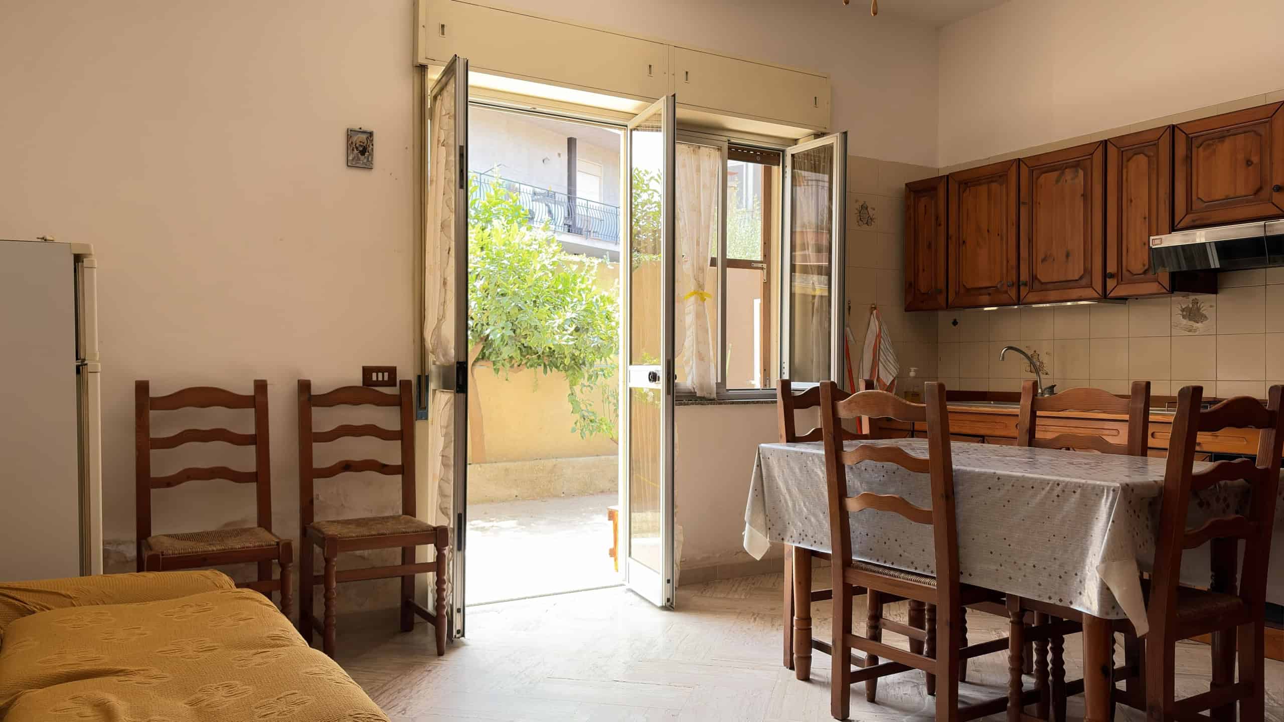 Interno di una cucina con tavolo, sedie in legno e porta finestra che dà su un balcone con piante verdi.