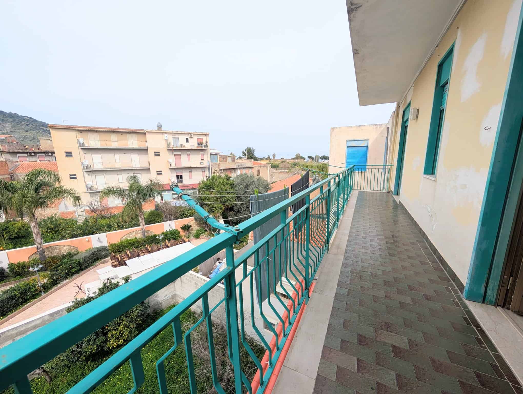 Balcone di un appartamento con ringhiera verde e vista su edifici e vegetazione circostante, in un giorno nuvoloso.