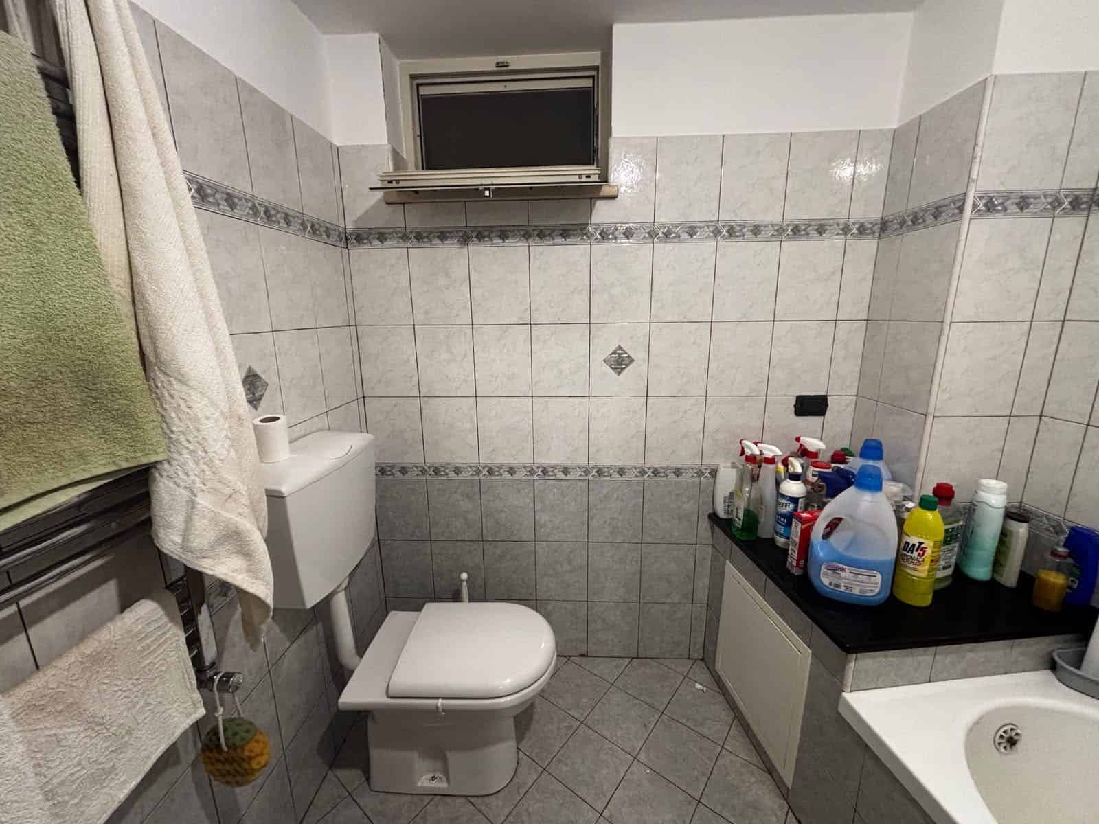 Bagno con sanitari, lavabo e prodotti per la pulizia, finestra alta, pareti rivestite di piastrelle grigie e bianche.