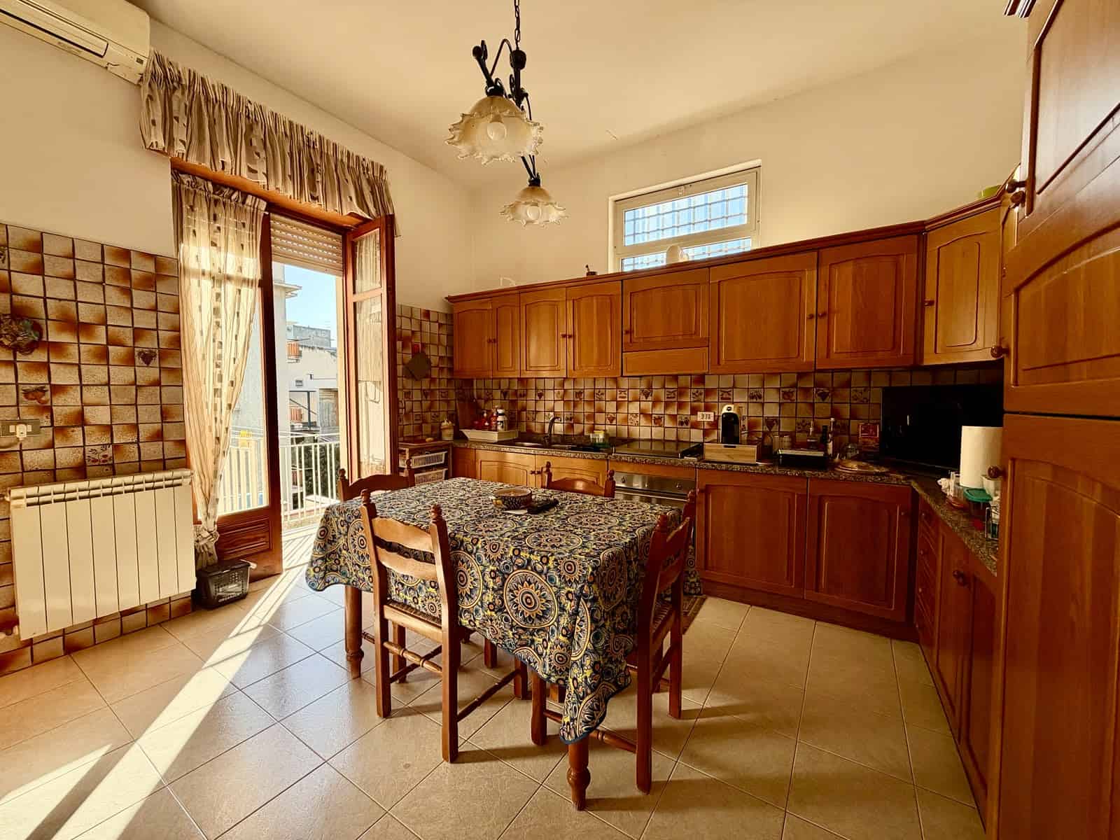 Cucina spaziosa con mobili in legno, tavolo con tovaglia e accessori vari, luce naturale da porta e finestra.