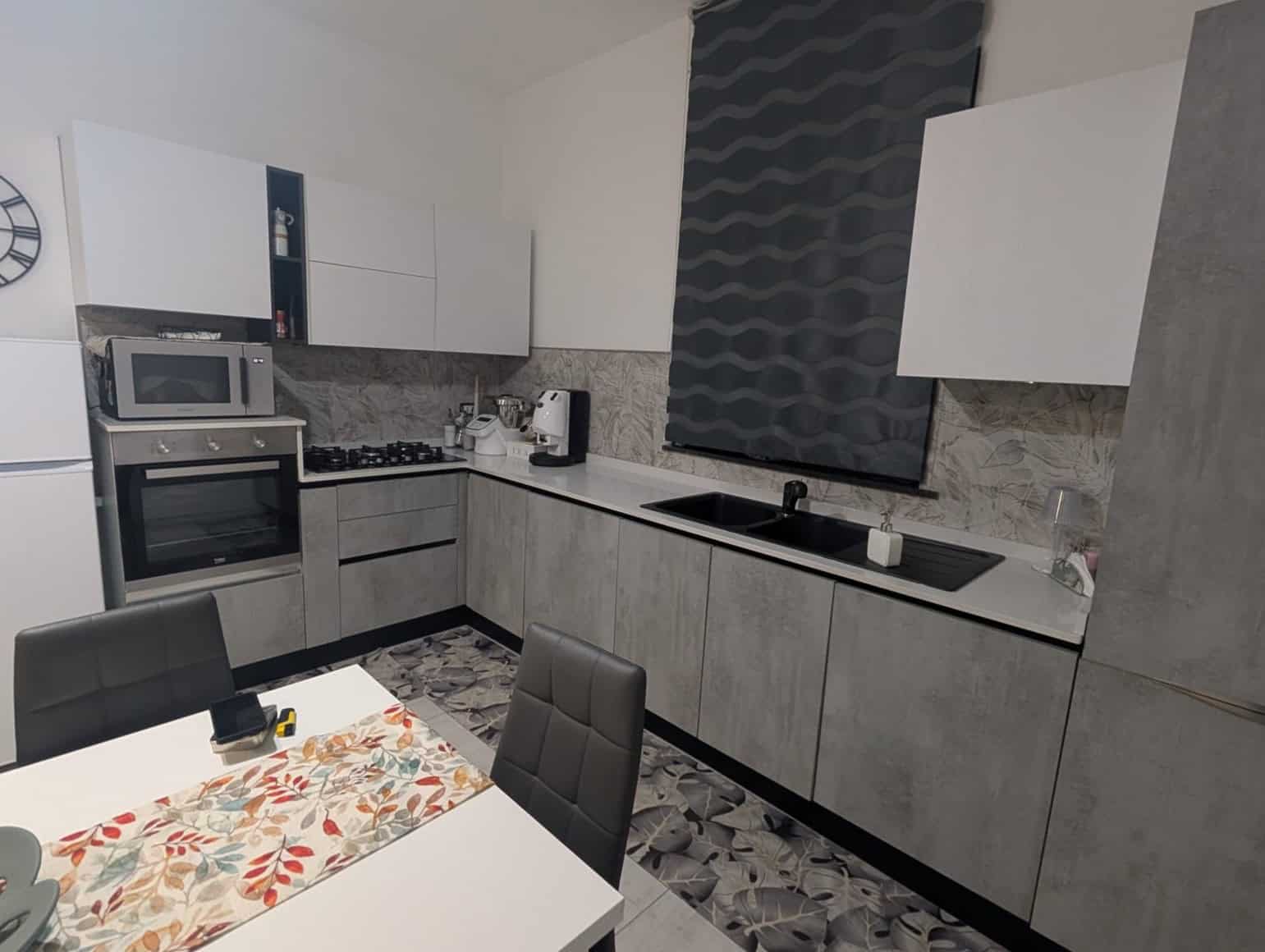 Un'ampia cucina moderna con elettrodomestici integrati, piano cottura a gas, lavello nero e armadietti bianchi e grigi, ideale per cucinare e socializzare in casa.